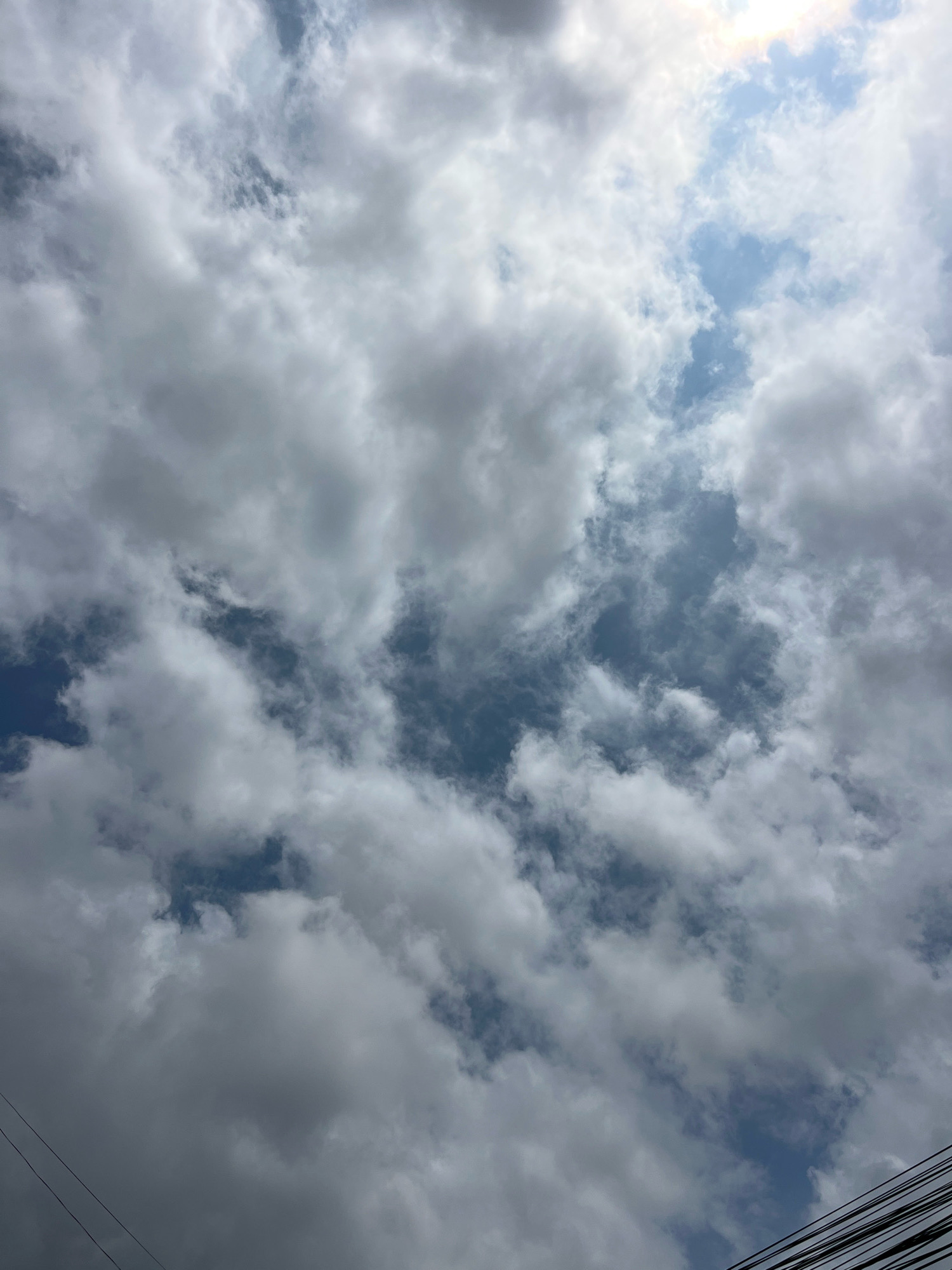 The image is a low-angle shot of a cloudy sky, with patches of blue visible through breaks in the clouds. The clouds are varied in texture and density, with some appearing wispy and others thick and grey. In the bottom left and bottom right corners of the image, several dark, thin wires are visible, suggesting the presence of power lines. The sky appears to be overcast with some areas of bright light peeking through, indicating it could be daytime. The scene is devoid of people, specific landmarks, or any discernible city elements, other than the power lines. No specific location within Denpasar, Indonesia can be identified from this image.
