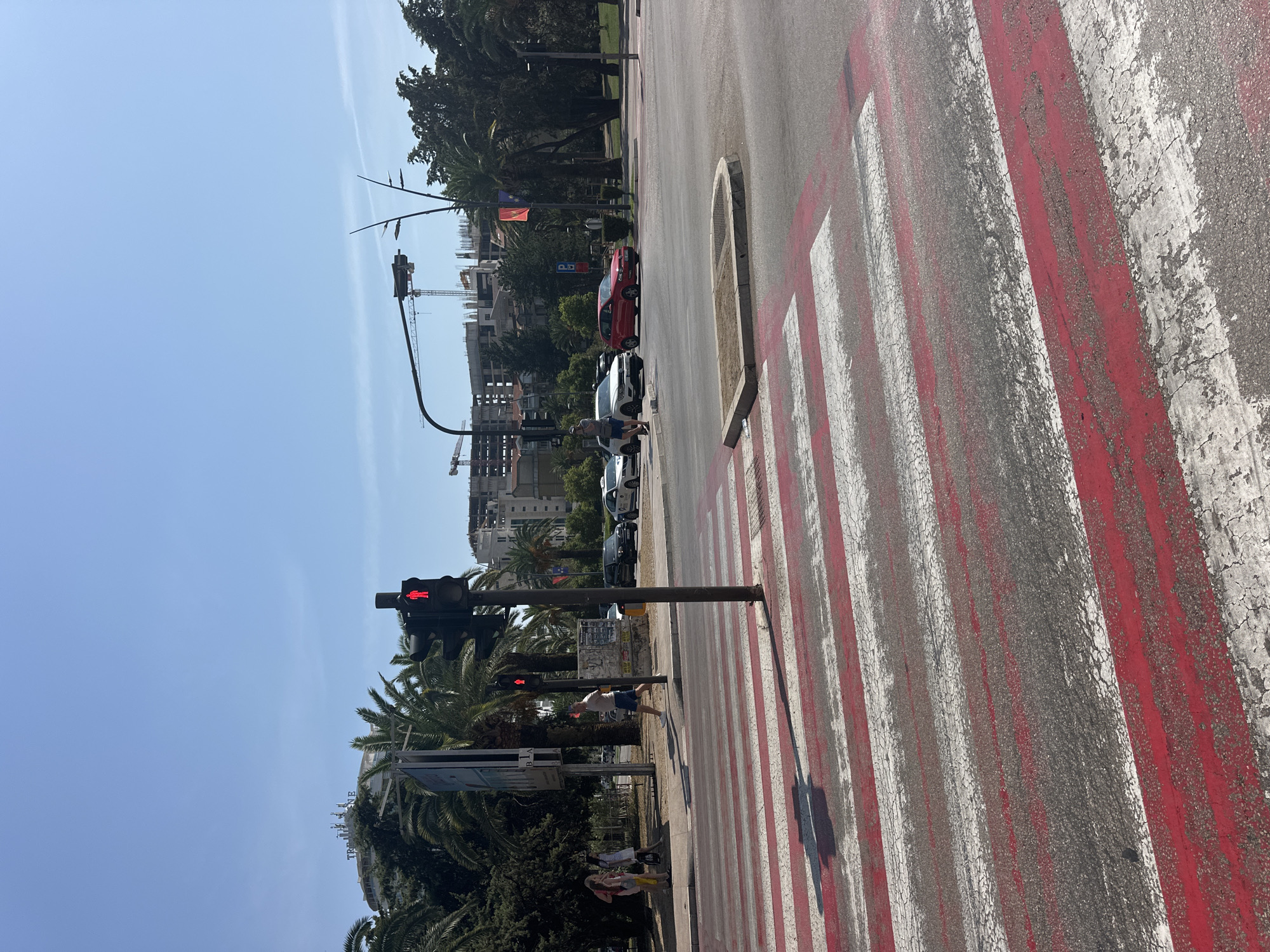 A bright, sunny day is depicted in this urban scene in Budva, Montenegro. A pedestrian crossing with faded red and white stripes is in the foreground. To the right of the crossing, a traffic light displays a red pedestrian signal, with a silhouette of a person walking. A man in a light-colored t-shirt and dark shorts is crossing the street, moving away from the camera. In the background, several cars are visible on the road, including a white SUV, a red car, and a white taxi. Trees and palm trees line the street. Construction cranes are visible in the distance, indicating ongoing development. The sky is clear and blue.