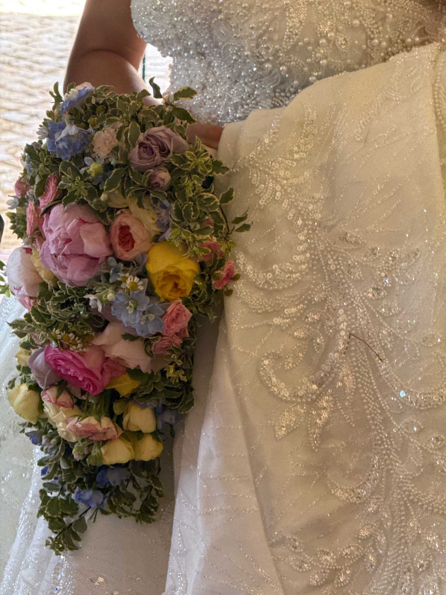 In Mafra, Portugal, a close-up shot captures a bride holding a cascading bouquet of flowers. The bouquet features a vibrant mix of pink peonies, yellow roses, blue delphiniums, and small white daisies, all nestled amongst lush greenery. The bride is wearing an ornate white wedding dress adorned with intricate beadwork and sequins. The background is blurred, suggesting an indoor setting with soft, ambient lighting.