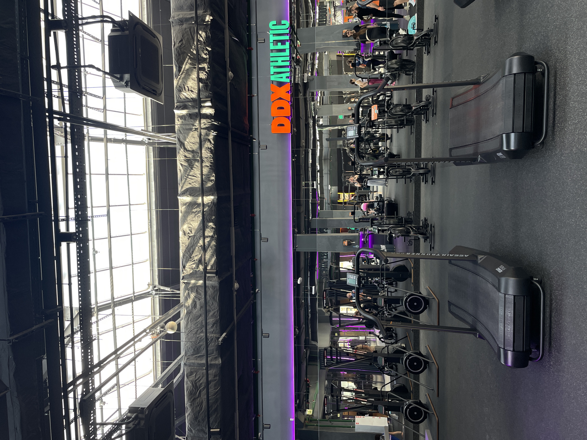 This is an interior shot of DDX Athletic fitness center in Kazan, Russia. A large, open space is filled with various exercise machines, including curved treadmills (Assault Runner Pro brand), stationary bikes, and other equipment. People are using some of the equipment. The walls are dark and lit with purple lights. A large window is visible, allowing natural light into the space. The gym's name, "DDX Athletic", is written in orange and green letters and is displayed on the wall. The image is rotated 90 degrees counter-clockwise.