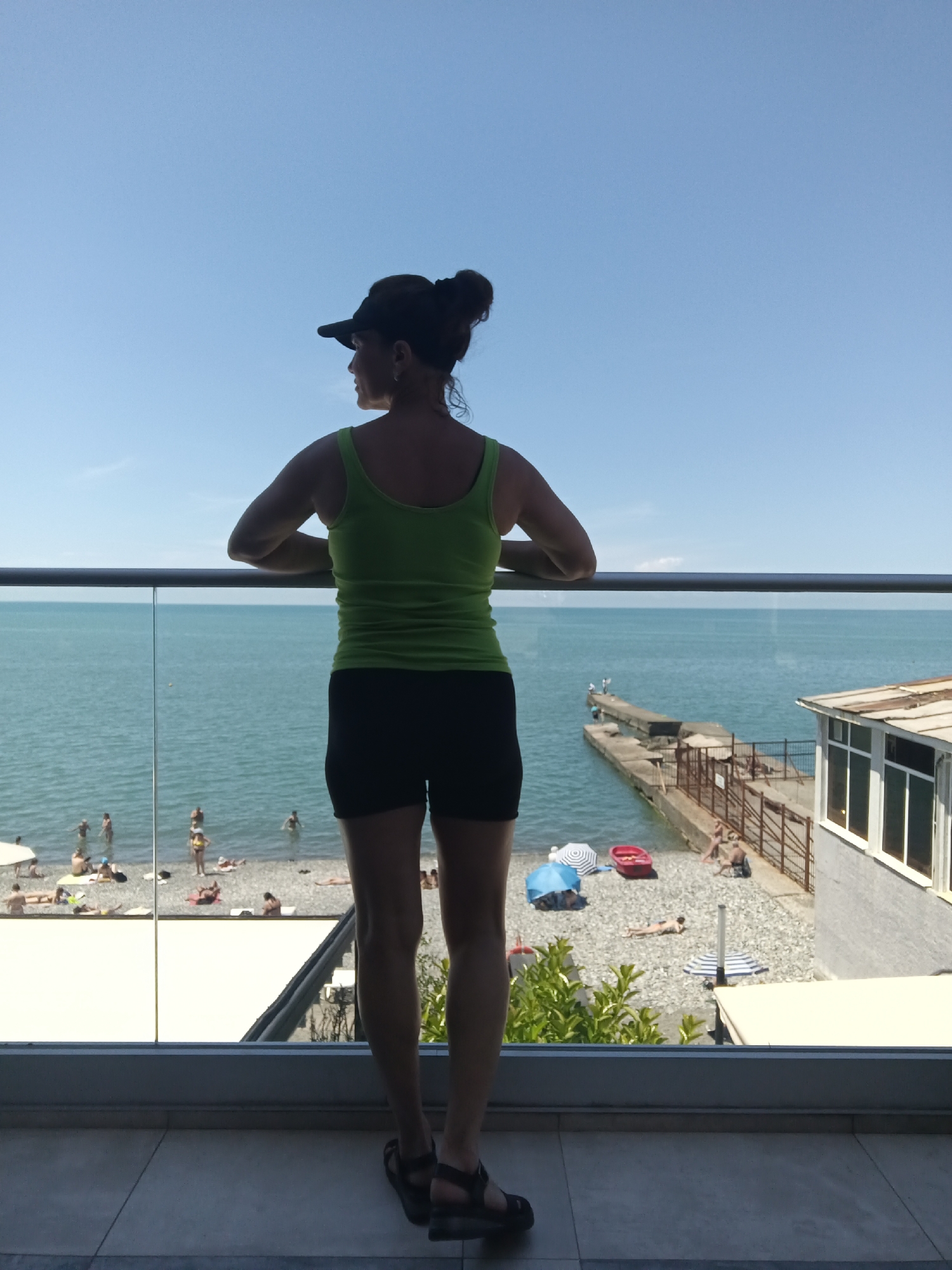 A woman stands on a balcony, facing away from the camera, looking out at the Black Sea in Sochi, Russia. She is wearing a green tank top, black shorts, a dark baseball cap, and sandals. The balcony has a glass railing. Below the balcony, a pebbled beach is visible with several people sunbathing and swimming in the turquoise water. A concrete pier extends into the sea, and a building with large windows is situated on the right side of the frame. The sky is clear and blue.