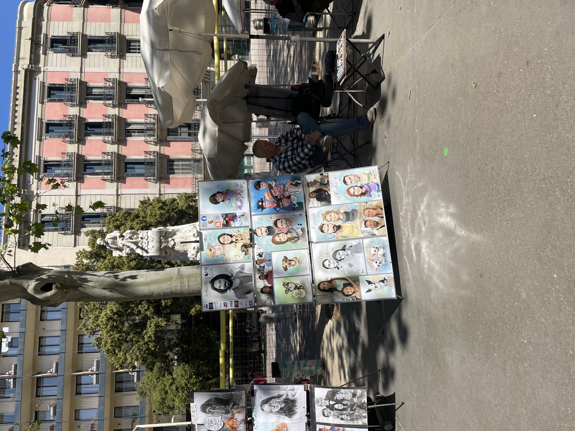 The image depicts an outdoor street scene, likely in a city square or pedestrian area in Barcelona, under bright sunshine. The main subject is a street artist, a man wearing a plaid shirt and jeans, seated on a folding chair under two large white umbrellas. He is positioned behind a large display stand showcasing a variety of his artworks.
The display is filled with numerous colorful caricatures and portraits. These include individual portraits, couples, families, and even pets, specifically dogs. The art style ranges from vibrant, exaggerated caricatures to more realistic black and white artistic portraits. Many of the caricatures feature cheerful and lively expressions.
Visible text on the display boards provides information such as "CARICATURAS" (Caricatures) and "PINTURAS" (Paintings), along with a price of "10€." Specific names like "Manuela" are also seen on some samples. The artist advertises various payment methods including "VIAL Bizum," "VISA," "AMERICAN EXPRESS," "Mastercard," "Paypal," and "Contactless." Social media icons for Facebook and Instagram, along with QR codes, are also present, suggesting the artist's online presence.
In the background, there is a tall building with a distinctive pink and white striped facade and numerous balconies. To the left, another building with a more modern, glass-fronted design is visible. Trees with green foliage and a white stone statue are situated between the buildings and the artist's setup. The ground is grey asphalt, and long shadows indicate a sunny day, likely midday or early afternoon.