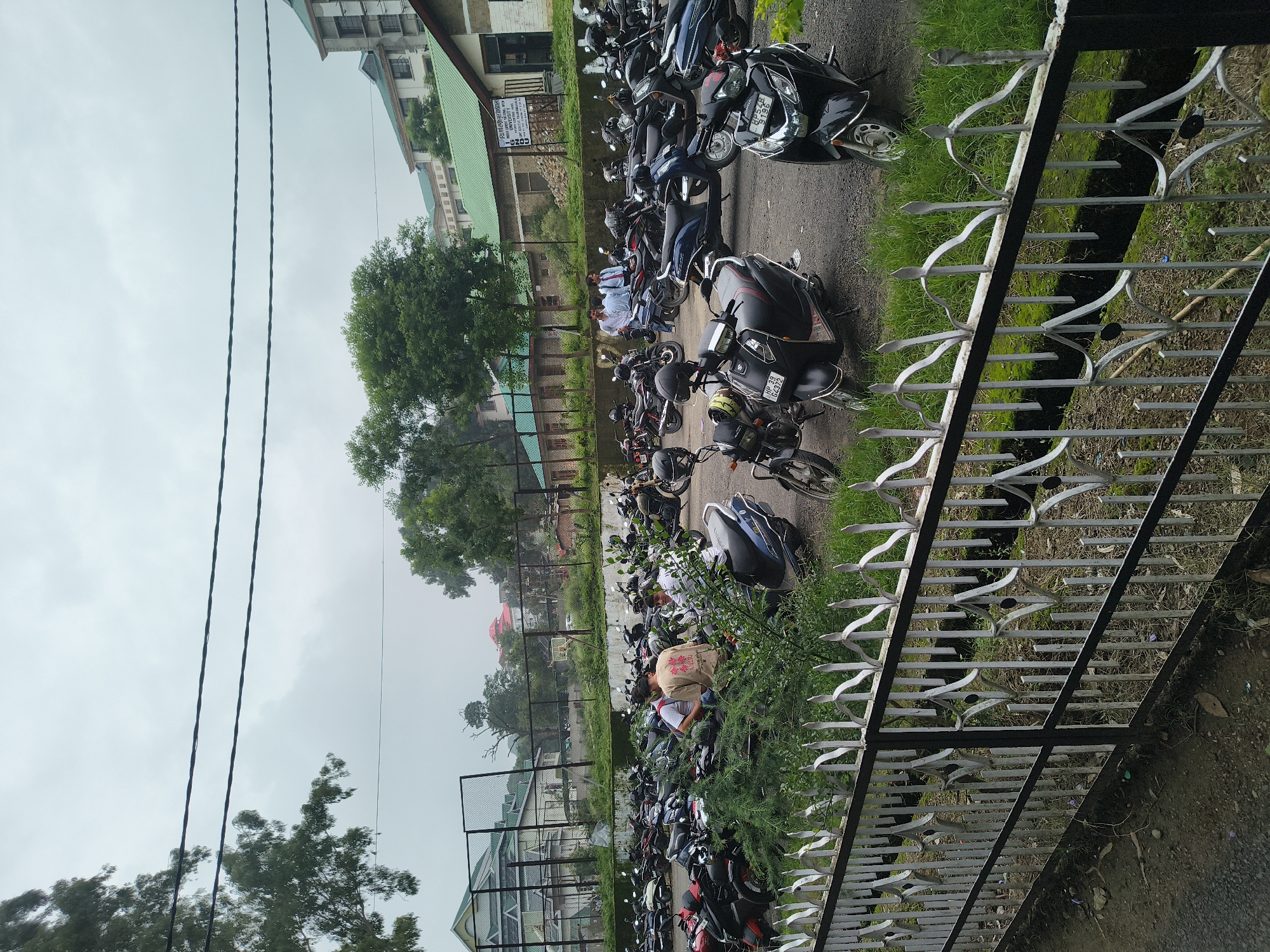 This is a photograph of a parking lot filled with motorcycles and scooters, likely at the Himachal Pradesh University in Shimla, India. The lot is paved and appears to be outdoors under an overcast sky. In the foreground, a decorative white metal fence with black accents runs across the bottom of the frame, with lush green grass and some low-lying shrubs visible behind it. Beyond the fence, numerous two-wheeled vehicles are parked in rows, filling much of the scene. Some individuals are standing or sitting near the motorcycles, appearing to be interacting or preparing to depart. In the background, several buildings can be seen, including structures with green roofs, typical of the architecture in the region. Trees are scattered throughout the area, contributing to a somewhat natural and academic environment. The overall impression is a busy day at a university campus parking facility.
