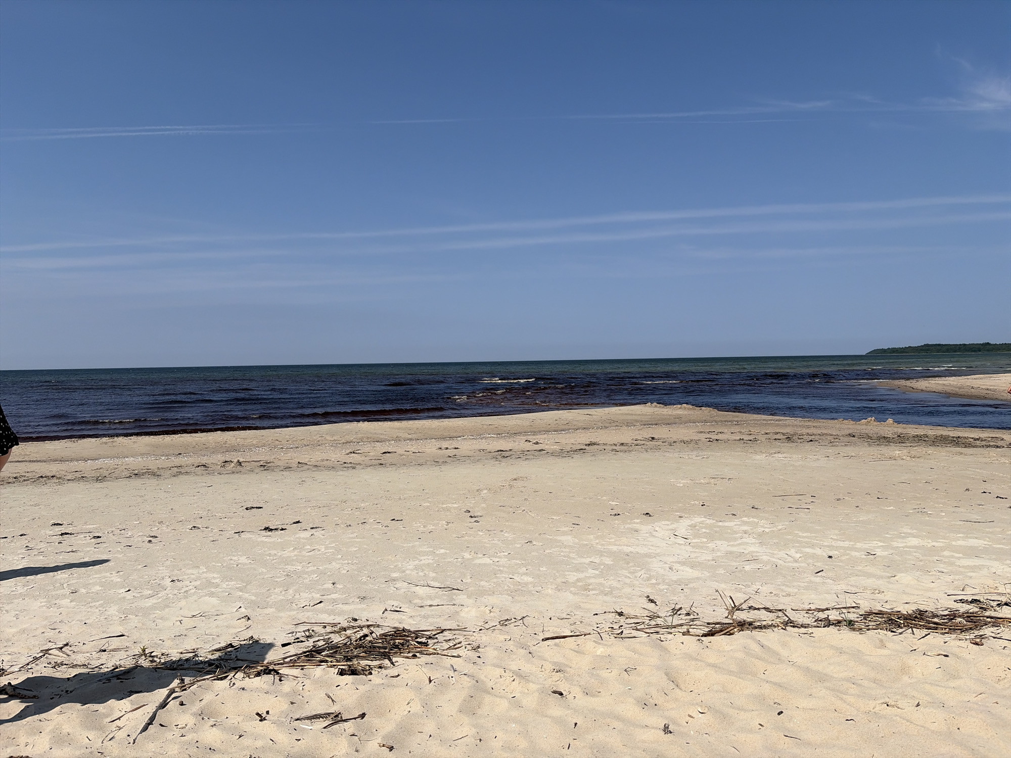 The image depicts a serene beach scene in Vääna-Jõesuu, Estonia. The foreground is dominated by a wide expanse of pale sand, scattered with dried seaweed and small branches. The sand exhibits ripples and shadows from unseen objects, suggesting a sunny day. In the midground, a shallow body of water with a darker hue meets the shore, with small waves gently lapping. Beyond this, the deeper ocean stretches to the horizon, its surface marked by subtle wave patterns. The sky above is a clear, bright blue, with faint, horizontal cloud streaks and a more prominent vapor trail from an aircraft stretching across the upper half of the frame. To the far right, a distant, tree-covered coastline is visible. On the far left edge of the image, a portion of a person's leg, clad in a black and white polka dot garment, is visible, casting a shadow onto the sand. The overall impression is one of natural tranquility and a peaceful coastal environment.