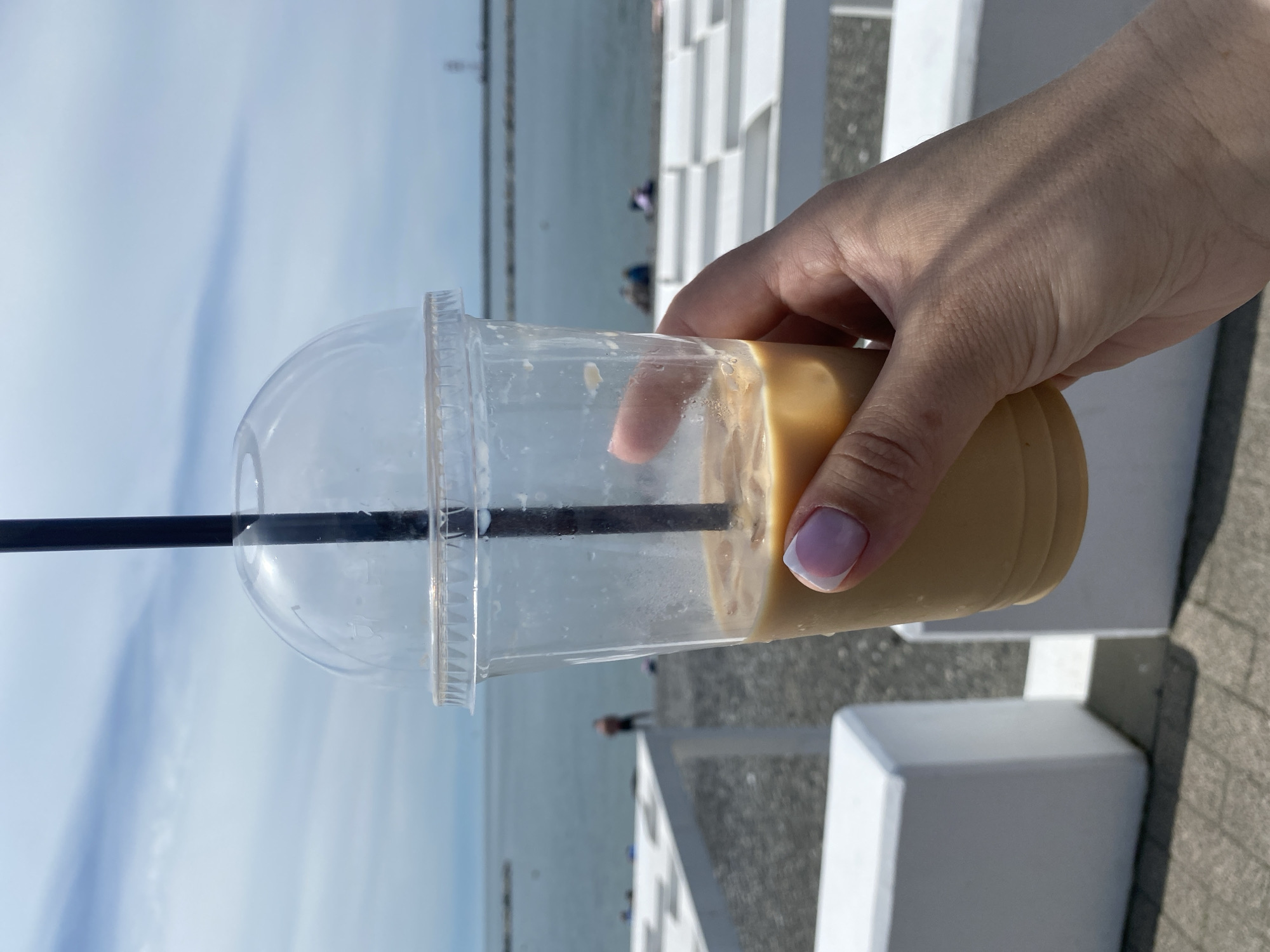 A medium shot captures a hand holding a clear plastic cup filled with a beige-colored beverage. A black straw extends from the cup's lid. The hand has pale skin and a neatly manicured nail with a light pink base and white tip.
The background reveals an outdoor scene. In the distance, a body of water stretches towards the horizon, with a pale blue sky overhead. On the right side of the frame, white, blocky structures or walls are visible, offering a contrast to the water and sky.
The image was taken in Sochi, Russia, as indicated by geolocation information. People can be seen indistinctly in the background near the sea.
