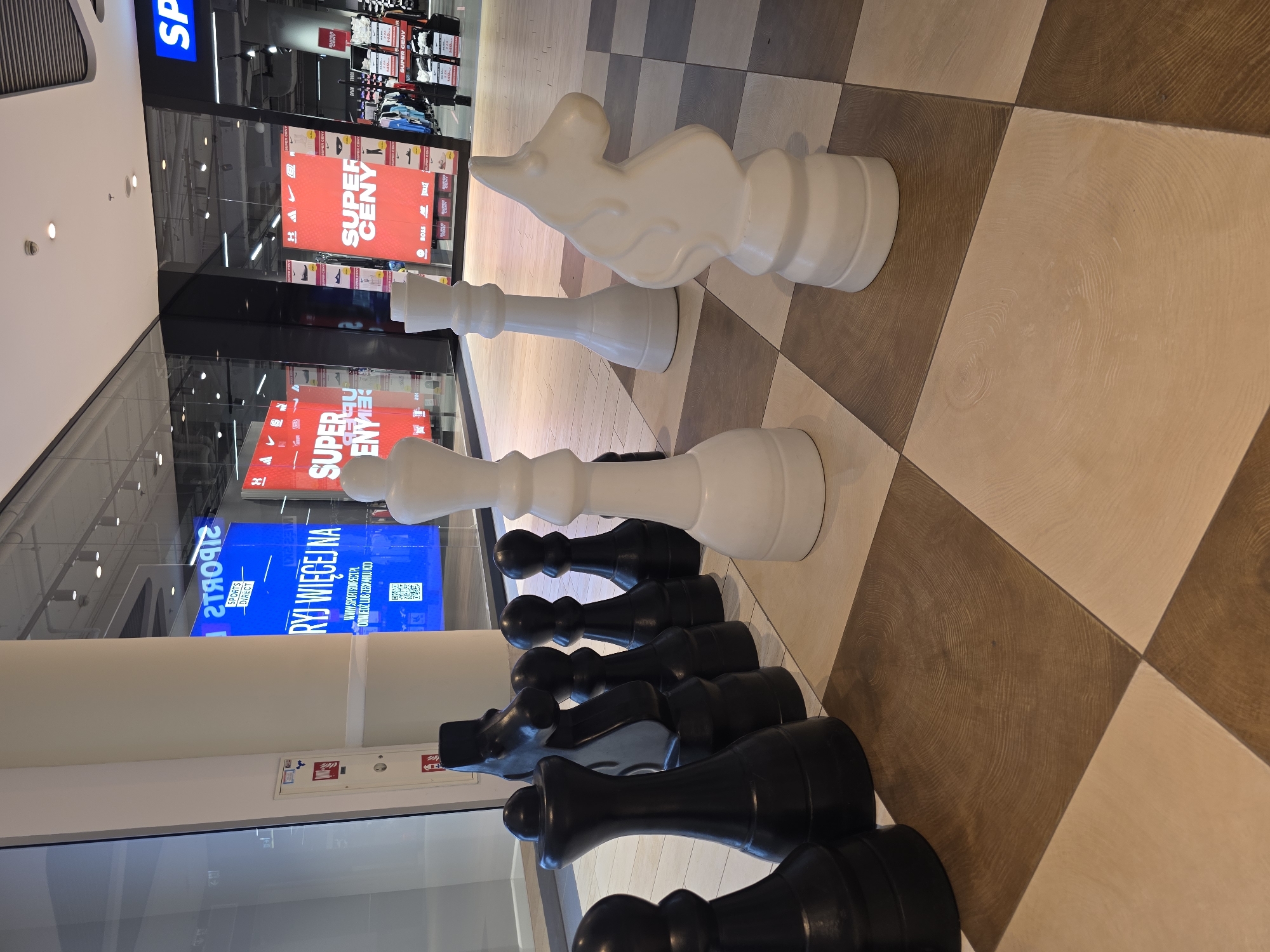 This media file captures a close-up view of oversized chess pieces, both white and black, arranged on a large checkered floor pattern that mimics a chessboard. The setting appears to be indoors, possibly a shopping mall or an exhibition space, given the reflections in the glass surfaces and the presence of retail signage in the background.
The white pieces visible are a king and a knight, positioned further back. In front of them, a line of black pieces, including several pawns and what looks like a rook, are lined up. The chess pieces are substantial in size, suggesting they are decorative or part of an installation. The floor is tiled with alternating light beige and darker brown squares, some featuring wood-grain-like patterns.
In the background, through a glass partition, there are signs advertising "SUPER CENY" (Super Prices in Polish) for a sports store, with brand logos like Nike and Adidas visible. One prominent blue digital display shows "SPORTS DIRECT" and Polish text, possibly "RYJ WIĘCEJ NA" (which translates to "Find more on"). The lighting suggests it is daytime, but the indoor environment makes it difficult to determine the time of day precisely. There are no people visible in the frame. The overall mood is still and somewhat monumental due to the scale of the chess pieces.