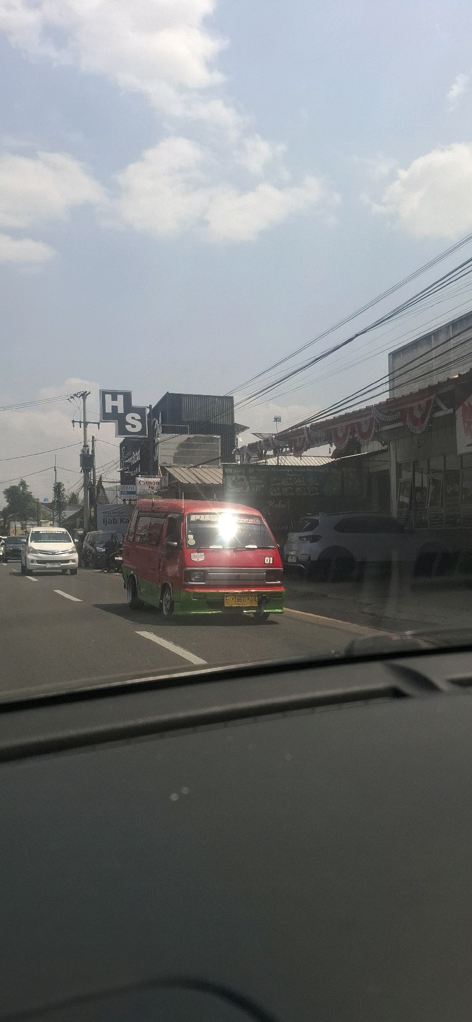 This is a vertical, eye-level shot of a street scene in Sukasari, Indonesia, on a bright, cloudy day. The foreground is dominated by the dark interior of a vehicle, likely the perspective of the photographer. In the midground, a red and green van, possibly a public transport vehicle, is the primary focus. It has the number "01" on its side and a license plate that appears to read "F 1960 PHM". The van's windshield is brightly reflecting the sun, obscuring some details of the interior. To the left of the van, a white sedan is visible, and behind it, several motorcycles are parked. The background shows a street lined with buildings, some with signage. One prominent sign has the letters "H S". Overhead, a network of power lines crisscrosses the sky. The overall impression is a typical urban street scene in Indonesia during daylight.