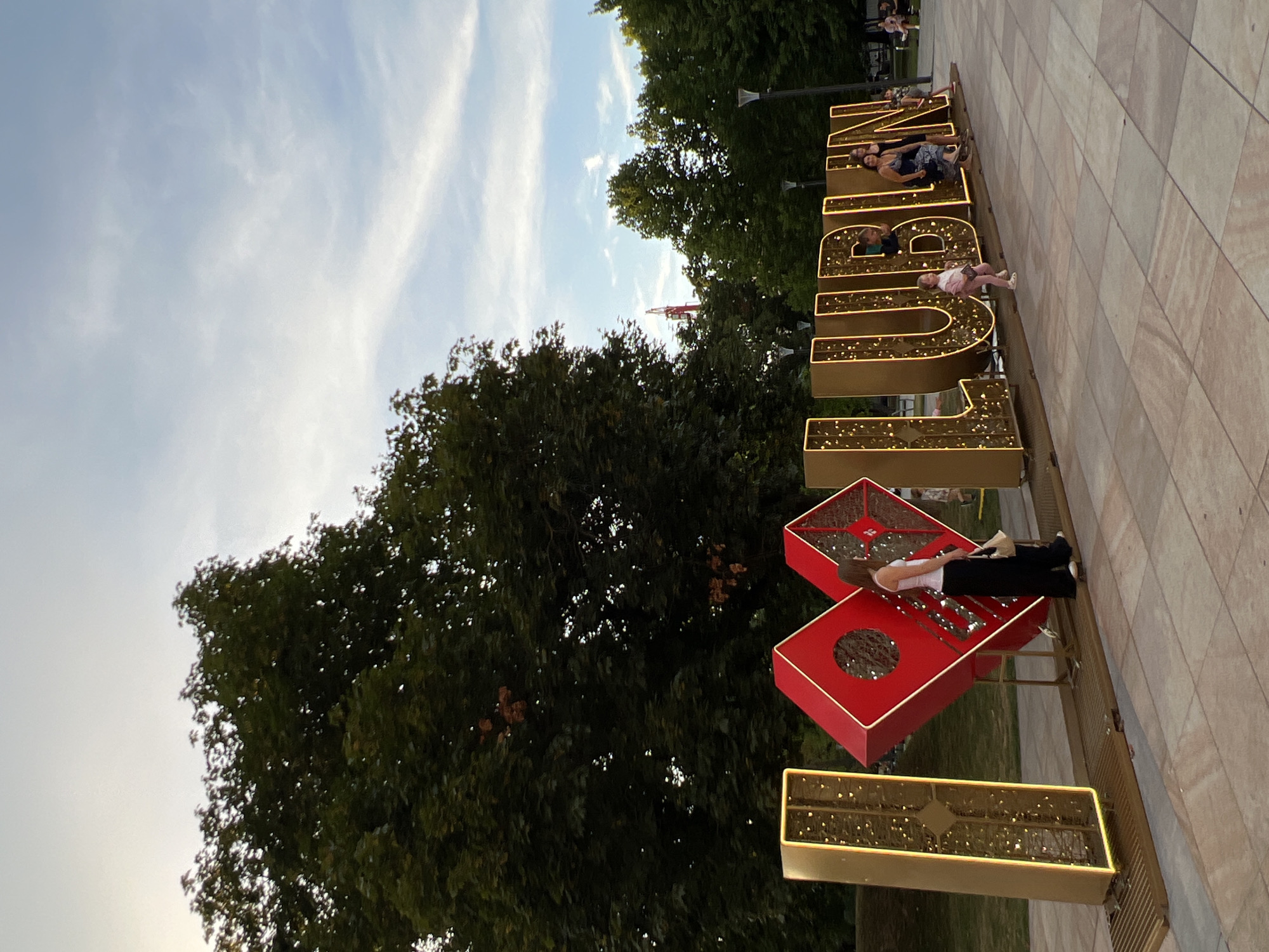 In an outdoor plaza paved with large, square tiles, a large, illuminated sign reads "I LOVE LUBLIN." The sign consists of individual, three-dimensional letters and symbols. The "I" is a tall, rectangular, golden structure with internal lights. Next to it, a large, red heart symbol with a diamond shape in the center is positioned at an angle. The letters "LUBLIN" are large, golden, and also illuminated with internal lights.
Several people are interacting with the sign. A woman in a white tank top and black pants is standing near the heart, looking at it. A young child in pink is walking in front of the "L" in "LUBLIN." Another woman in a blue and white patterned dress is sitting on the base of the "B" in "LUBLIN," with another person next to her. In the background, more people are visible in the distance, some sitting on benches.
The environment surrounding the plaza includes a grassy area with trees and a pathway. The sky above is a pale blue with scattered white clouds. In the far background, a red crane is visible above the trees, suggesting a construction site or landmark in the city. The lighting suggests it is either late afternoon or early evening.