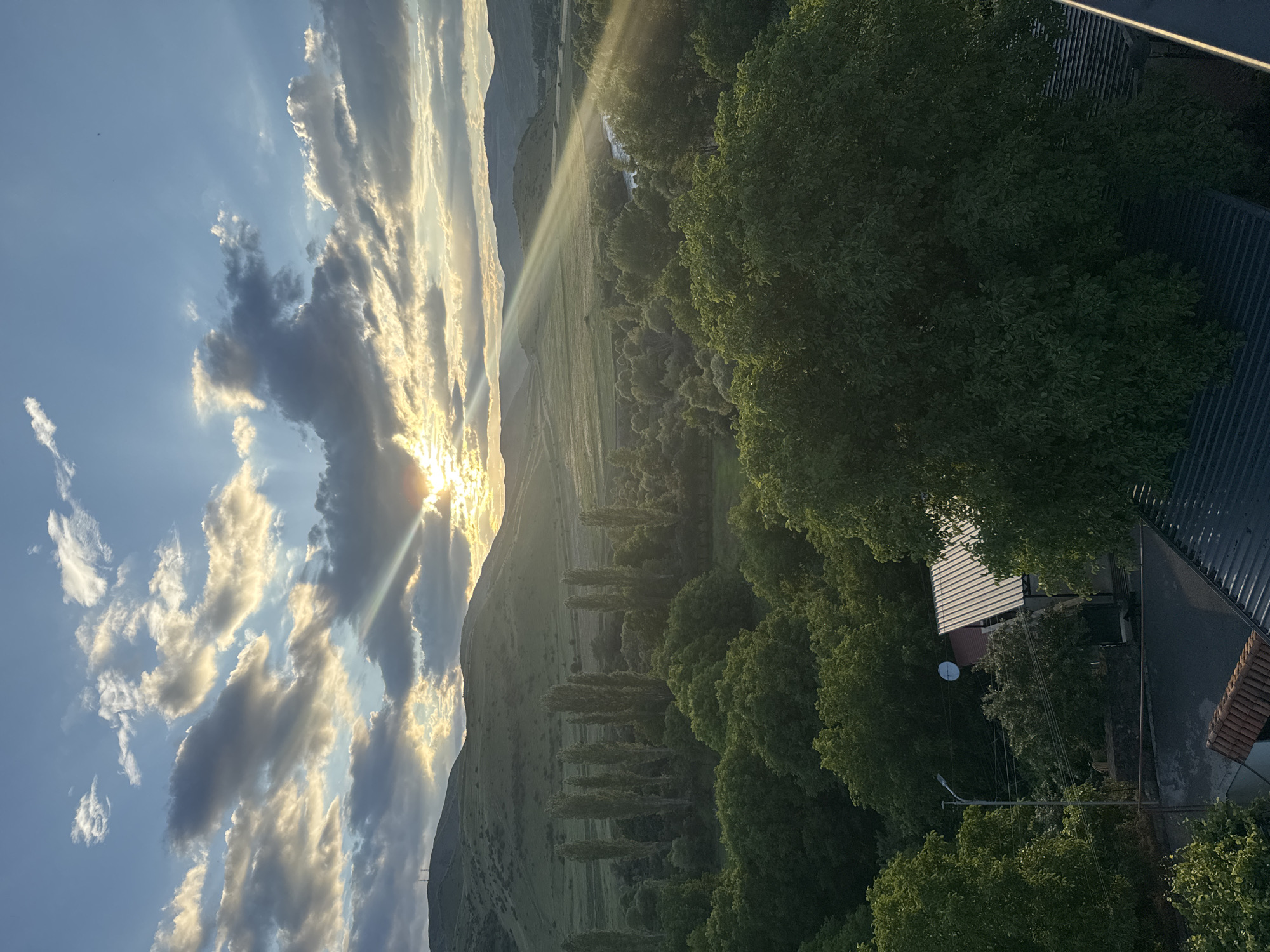 The image depicts a scenic view of the city of Aspindza, Georgia, during what appears to be sunset or sunrise. The sky is dramatic with a mix of dark and light clouds, with sunbeams piercing through, creating a ray effect. In the foreground, there are rooftops of houses, suggesting an elevated viewpoint. Beyond the houses, a lush green landscape unfolds, characterized by dense trees and rolling hills in the background. A river or body of water can be seen winding through the valley. The overall impression is one of natural beauty and tranquility. There are no people or specific actions visible in the image.