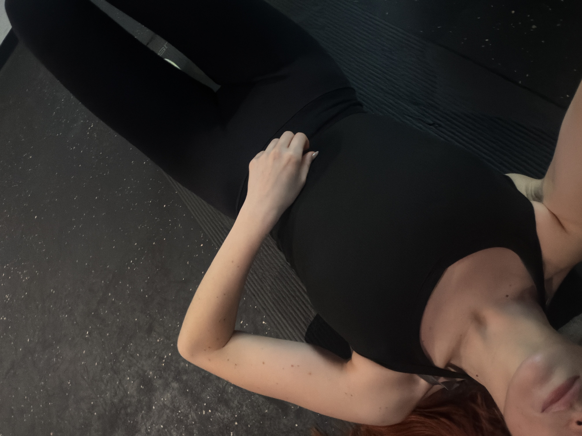 A woman is lying on her back on a gray and black workout mat, wearing black leggings and a black tank top. Her right arm is extended along her side, with her hand resting on her abdomen. She has red hair and fair skin, and is lying on a gray floor, suggesting a gym environment. The image does not provide any location information to identify Paris.