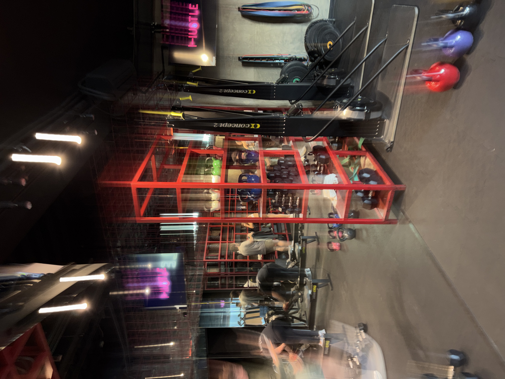 The image captures a workout space in Mexico City, Mexico. The space features rows of equipment, including several "Concept 2" rowing machines. Weight racks are stocked with dumbbells and kettlebells. A few individuals are engaged in exercising on stationary equipment. A sign with magenta lettering is partially visible. The overall lighting is a mix of bright fluorescent and neon.