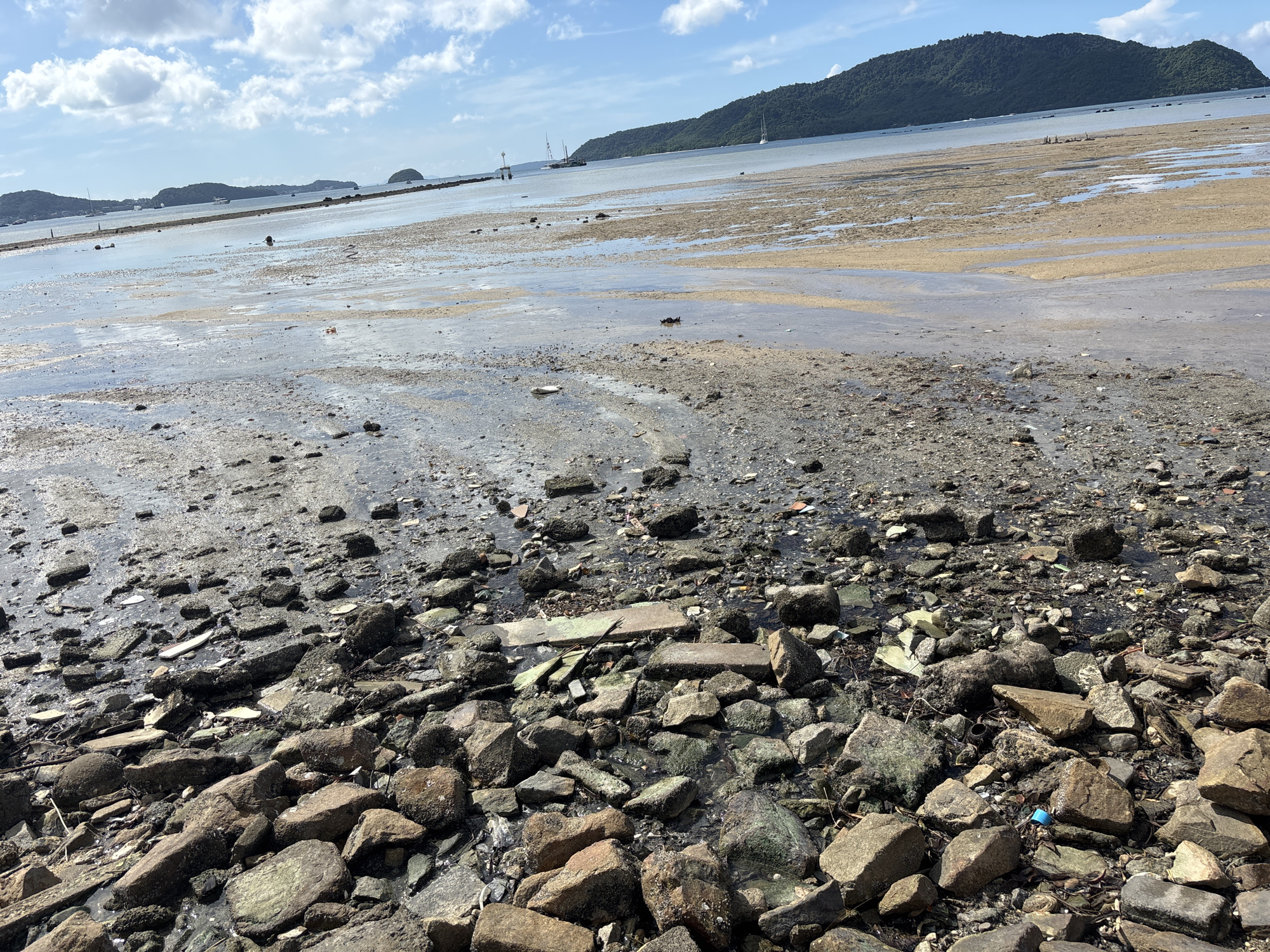 This is a wide-angle, low-angle shot of a rocky beach at low tide in Rawai, Thailand. The foreground is dominated by a dense collection of various-sized rocks and stones, some wet and reflecting the light. Interspersed among the rocks are fragments of debris, including what appear to be broken pieces of concrete or tile. A small blue plastic bottle cap is visible among the rocks in the lower right quadrant. The middle ground shows a muddy, wet sandbar dotted with shallow pools of water and more scattered debris. In the background, the sea extends towards a distant, tree-covered island. Several sailboats and other vessels are visible on the water, and a low, man-made barrier stretches across the sea in the distance, possibly a breakwater or pier. The sky is blue with scattered white clouds. No people are clearly visible in the frame.