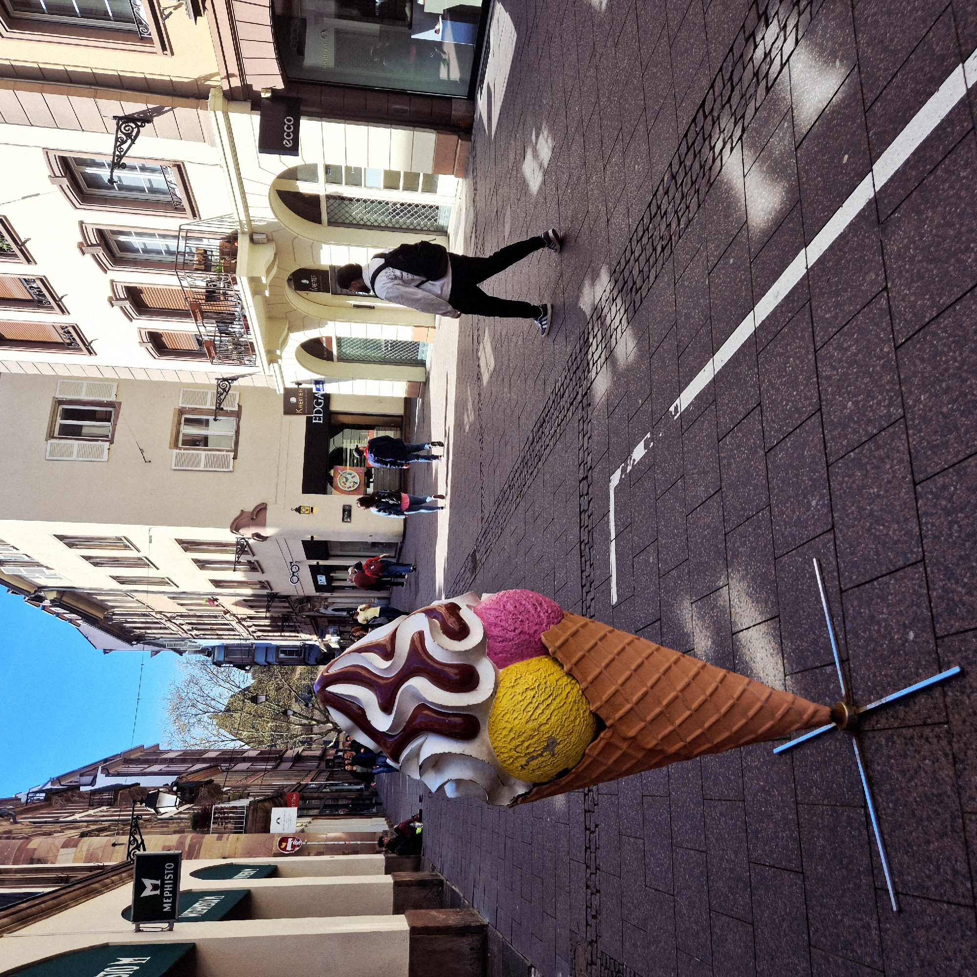The image shows a pedestrian street in Strasbourg, France. The street is lined with buildings, a large ice cream cone sculpture, and several people. A person wearing a white hoodie and black pants with a backpack is walking away from the camera. There are other people walking behind them. The street is paved with brown bricks. A white line is painted on the street. The ice cream cone has pink, yellow, and white scoops.