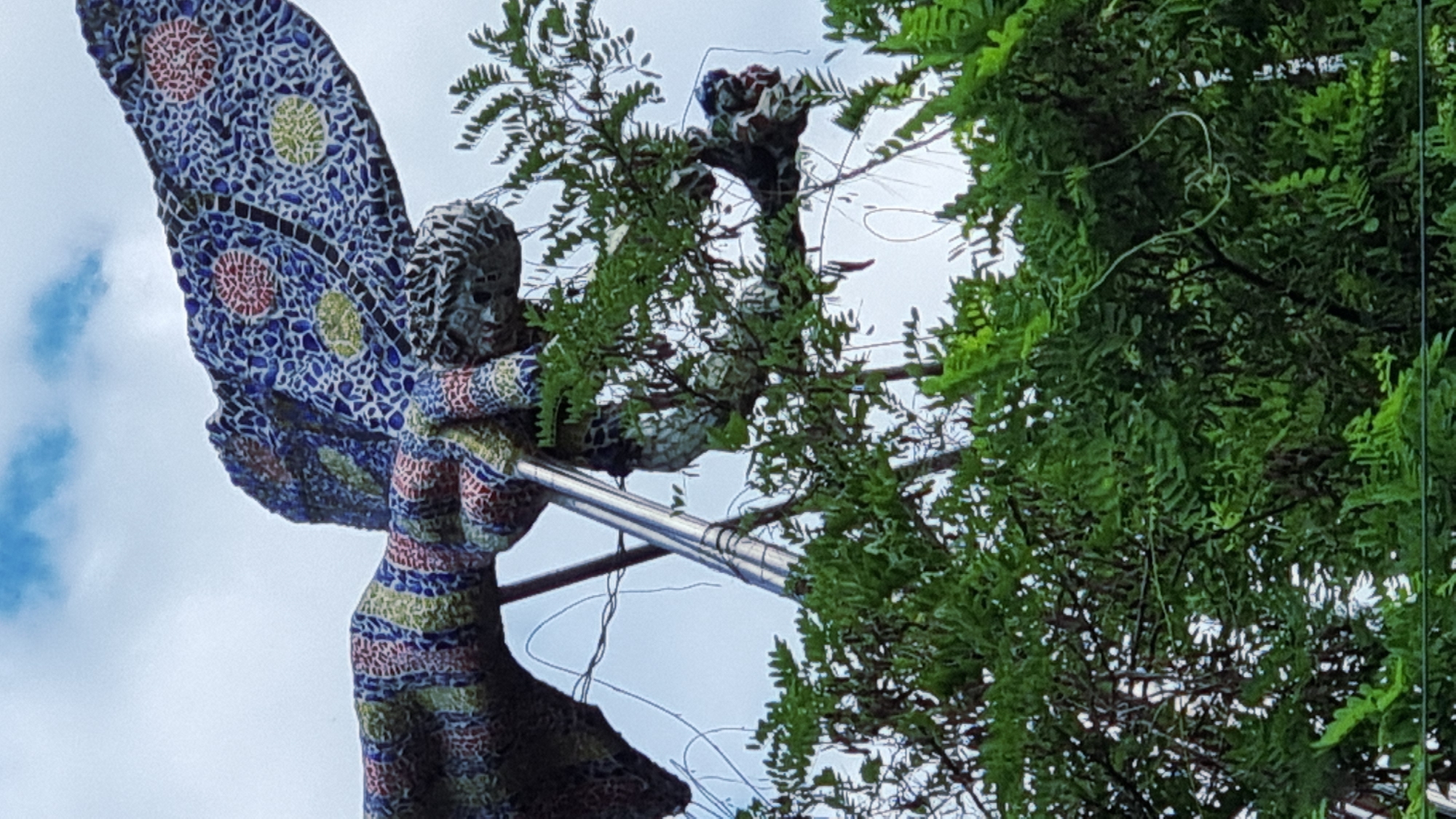 A mosaic sculpture of a dragonfly is positioned high up, partially obscured by lush green foliage. The sculpture is vividly decorated with a mosaic of small, colorful tiles in patterns of blue, red, yellow, and green. Its wings are spread, showcasing intricate circular designs. A metallic rod or pole supports the sculpture from below, running vertically through the frame. The background consists of a pale, cloudy sky. The lower portion of the image is dominated by the dense, leafy branches of trees. The scene is situated in Sant Joan Les Fonts, Spain.