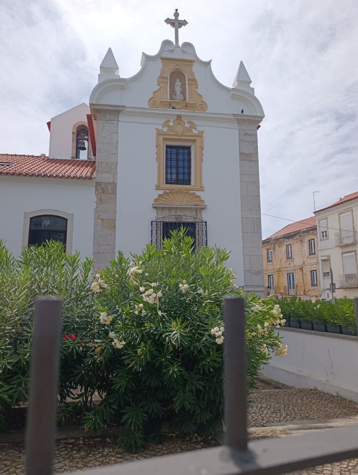The image shows the facade of a white church with a bell tower to the left, adorned with a red-tiled roof. The church features ornate architectural details, including a cross at its apex, a statue in a niche, and a decorative window. In the foreground, lush green bushes with white flowers partially obscure the view of the church and a paved pathway. To the right of the church, residential buildings with red-tiled roofs and balconies are visible. The sky is overcast with clouds. Based on the architectural style and the surrounding buildings, the location is likely Setúbal, Portugal, specifically the Church of São Lourenço or a similar historic religious building in the city. There are no people or animals visible in the image.