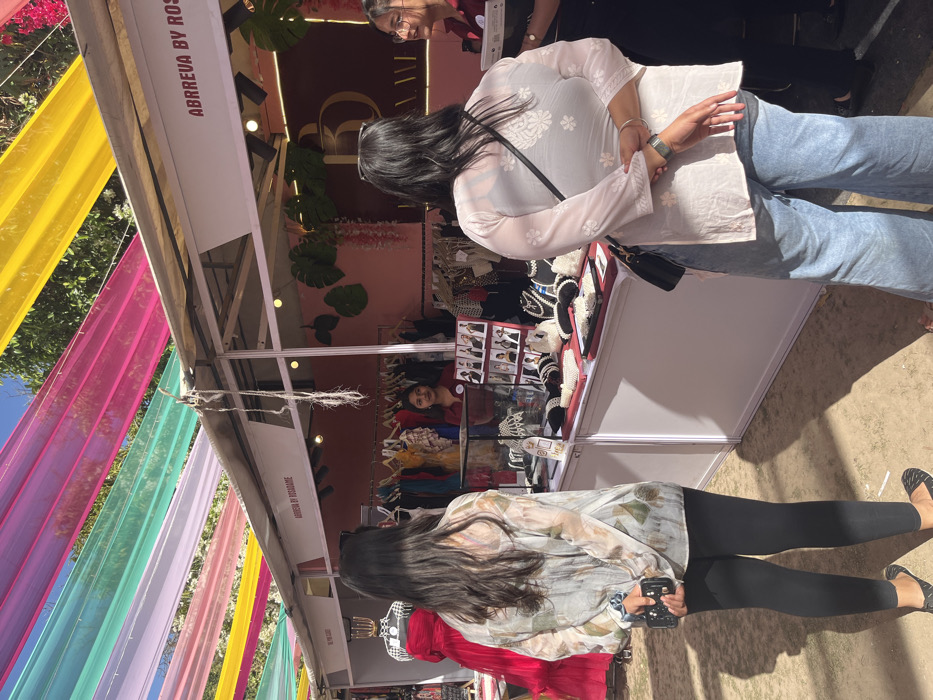 The image shows a market stall in Chandigarh, India, located at the coordinates 30.764 latitude and 76.798 longitude. A woman in a white floral blouse and black pants is looking at the display of necklaces and jewelry on a table inside the stall. She is holding a cell phone in her left hand. Another woman, wearing a white embroidered top and blue jeans, is standing behind her, looking at the jewelry. There is a third person in the background, a young woman wearing a red top and holding a tray of jewelry. The stall is decorated with pink and white fabric and has a sign that reads "ABBREVA BY ROS". The stall is situated under a canopy of colorful fabric that is strung overhead. The ground is dirt. The sun is shining brightly.