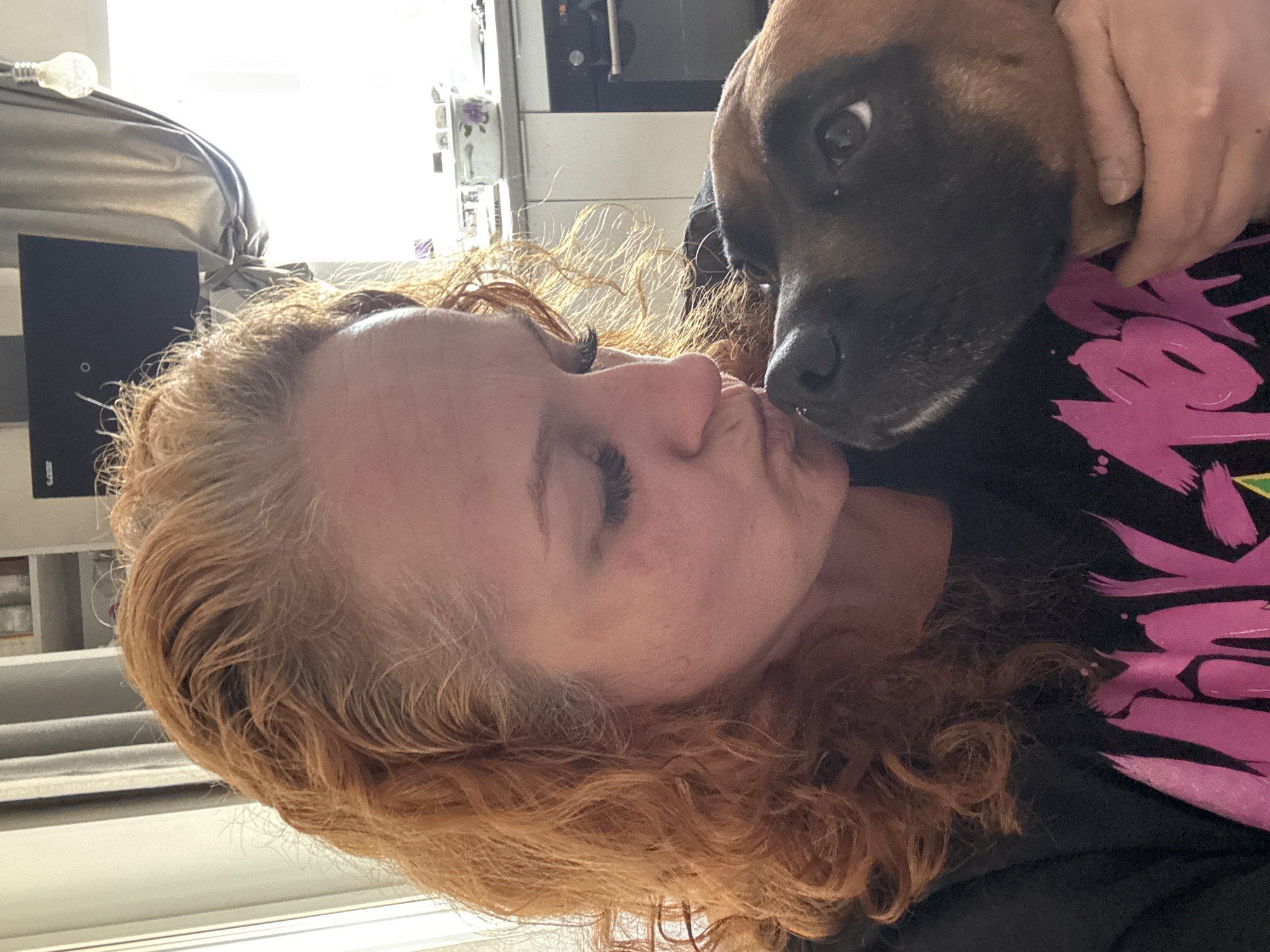 The image shows a woman with curly red hair holding a brown and black dog close to her face, appearing to kiss it. The woman has her eyes closed. They are indoors. In the background, there is a window with blinds and a shelf. The location is unknown.