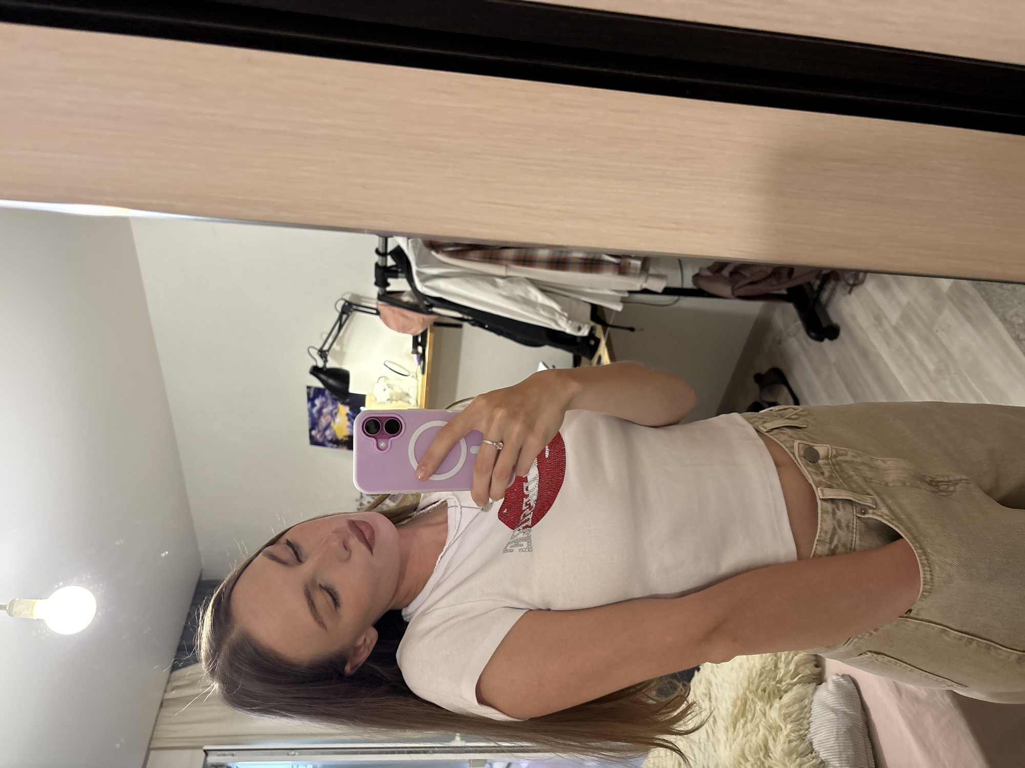 A young adult female is depicted in a mirror selfie taken within a residential room in Perm, Russia. The individual has long brown hair, fair skin, and is wearing a white short-sleeved crop top featuring a stylized design of red lips with the word "BABE" in silver/white letters, and light beige loose-fitting pants. Her head is tilted downwards with eyes closed or looking away from the camera. She holds a purple smartphone with a prominent circular magnetic ring in her left hand, wearing a silver ring on her ring finger. Her right hand is placed inside the pocket of her pants. The background, reflected in the mirror, reveals a white ceiling with an exposed light bulb, white walls, and a window with white curtains on the left. To the right, a light wood-paneled wardrobe is visible. Behind the individual, a desk or shelf area is equipped with a black articulated desk lamp, a framed purple and yellow artwork, and various small items. A clothes rack behind the desk holds multiple garments, including a light-colored jacket, a plaid shirt, and a pink cap. The floor is light-colored wood or laminate, and a furry rug and a corduroy pillow are visible on a pink surface to the left of the individual.