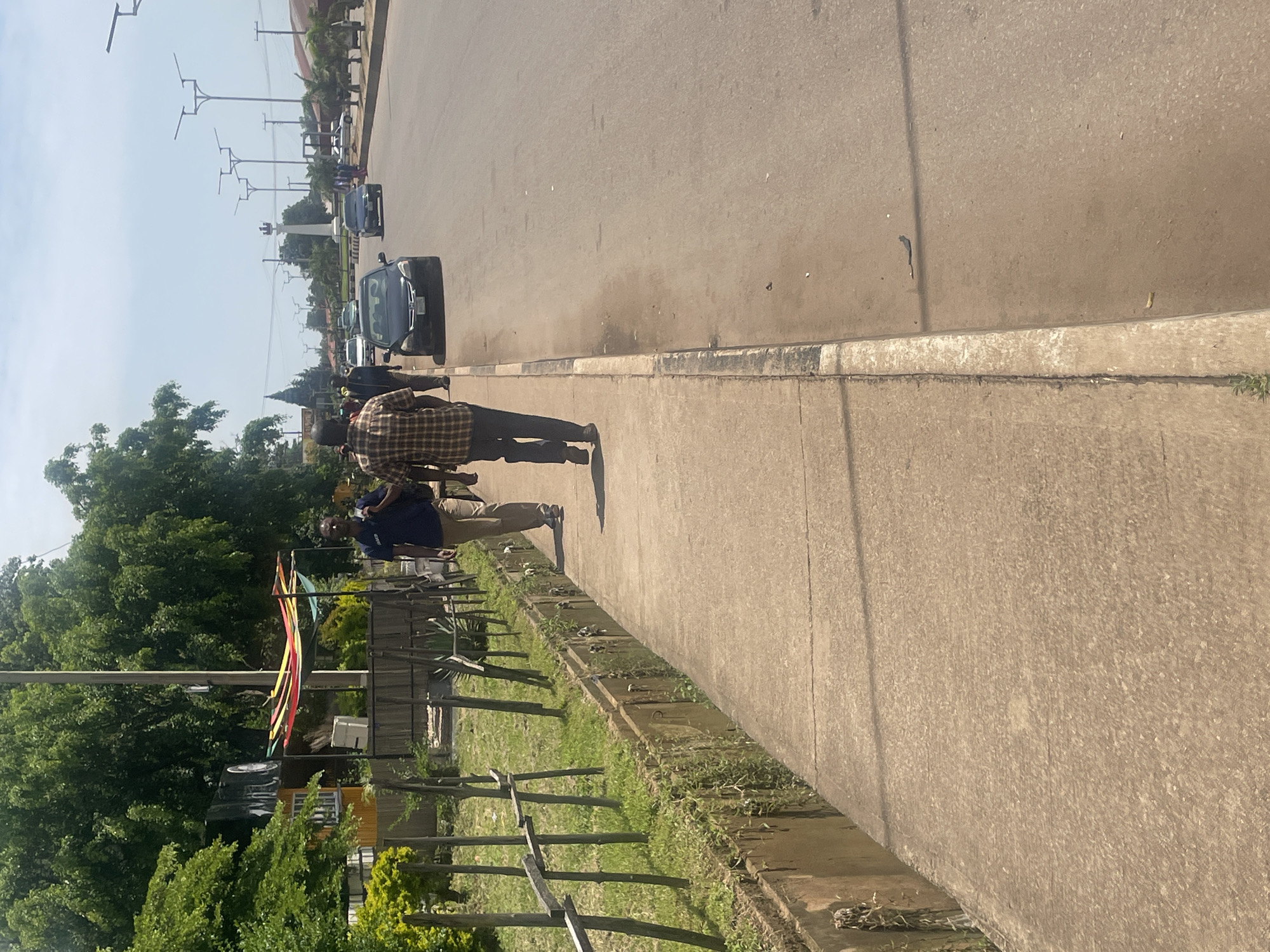 Two men are walking on a concrete sidewalk next to a road in Gura Top, Nigeria. The man in the foreground is wearing a plaid shirt and dark pants. The man behind him is wearing a dark blue shirt and khaki pants. The road has at least two lanes and several cars are visible. The sidewalk is bordered by a concrete ditch and then a grassy area with wooden posts. In the background, there are trees, buildings, and streetlights. The sky is clear and blue.