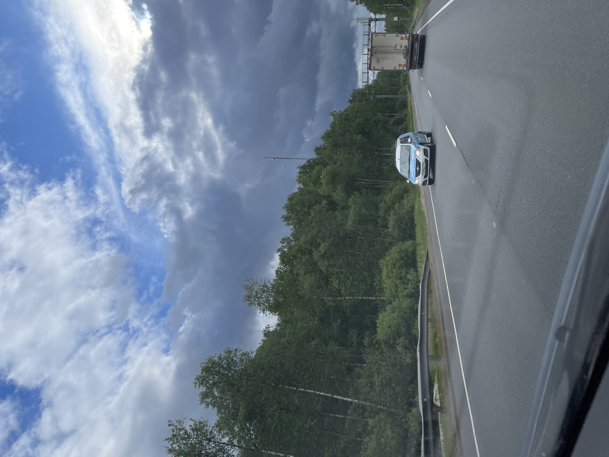 The image shows a highway near Novyi Byt, Russia. A light blue and white car and a truck are traveling on the wet asphalt road, alongside a dense forest under a cloudy sky.