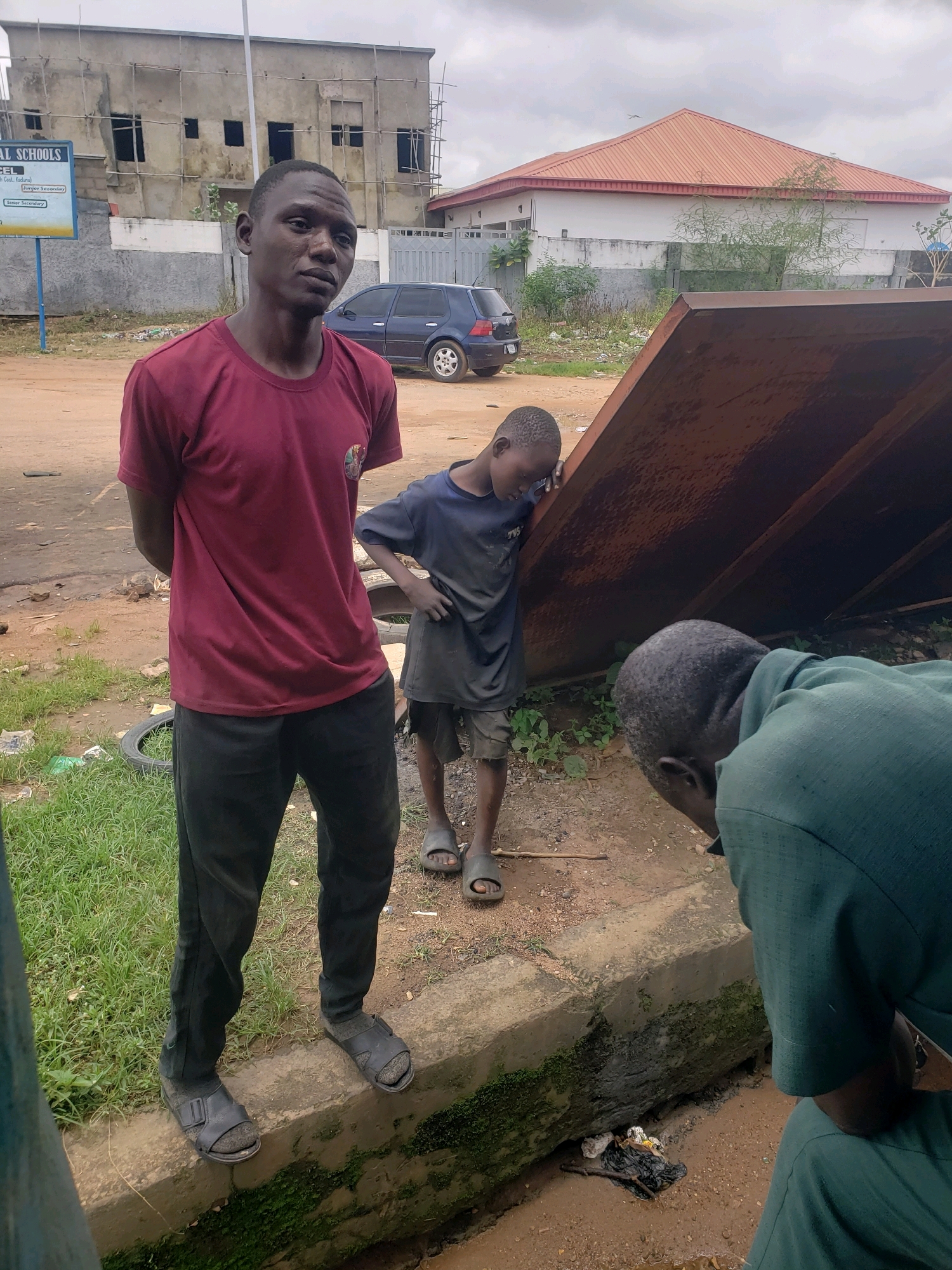 The image depicts an outdoor scene in Sabon Tasha, Nigeria, with a focus on three individuals and their surroundings. In the foreground left, a man with dark skin and short hair stands facing the camera, wearing a maroon t-shirt and black pants with black sandals. His arms are behind his back. To his right and slightly behind, a young boy with dark skin and short hair, wearing a dark t-shirt and shorts with sandals, leans his head against a large, rusty, angled metal surface. In the lower right corner, another individual, seen from the back and side, wears a green jacket and is bending over, looking down at a ditch filled with water and debris. The background shows a partially constructed building with scaffolding, a blue car parked on a dirt road, and a white building with a red roof. The ground is a mix of dirt and grass, and there is a concrete curb along the ditch. The sky is overcast.