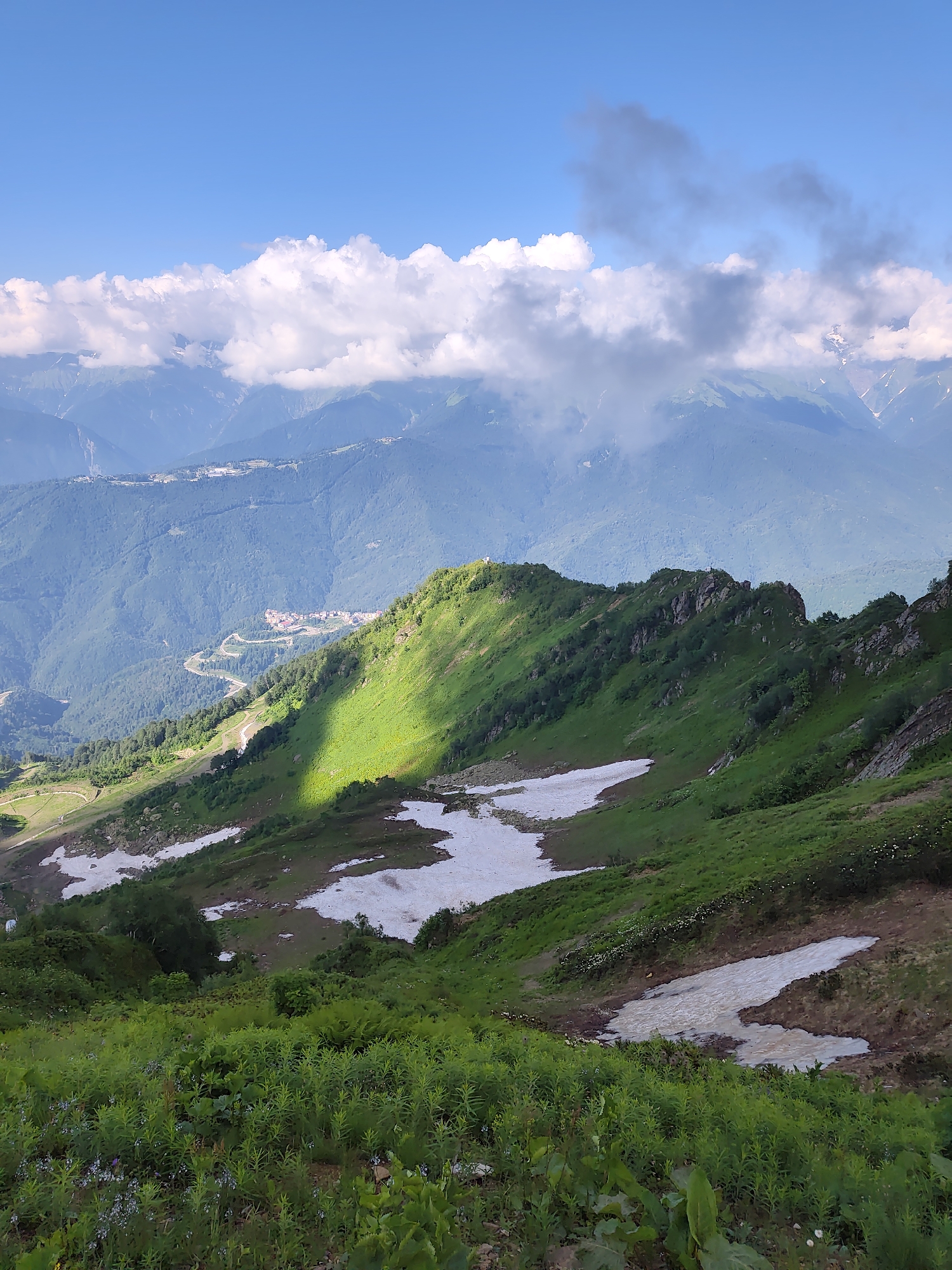 This image captures a scenic mountain landscape in Estosadok, Russia. The foreground features lush green vegetation with scattered patches of snow. A steep, verdant mountainside dominates the midground, with a distinct shadow cast by a sunlit ridge. In the distance, rolling hills covered in dense forest lead to higher, mist-shrouded peaks. A winding road is visible on one of the lower slopes, with buildings nestled further back. The sky is a vibrant blue, punctuated by a band of white clouds above the distant mountains. The overall scene depicts a natural environment during a time of day when sunlight dramatically illuminates parts of the landscape.