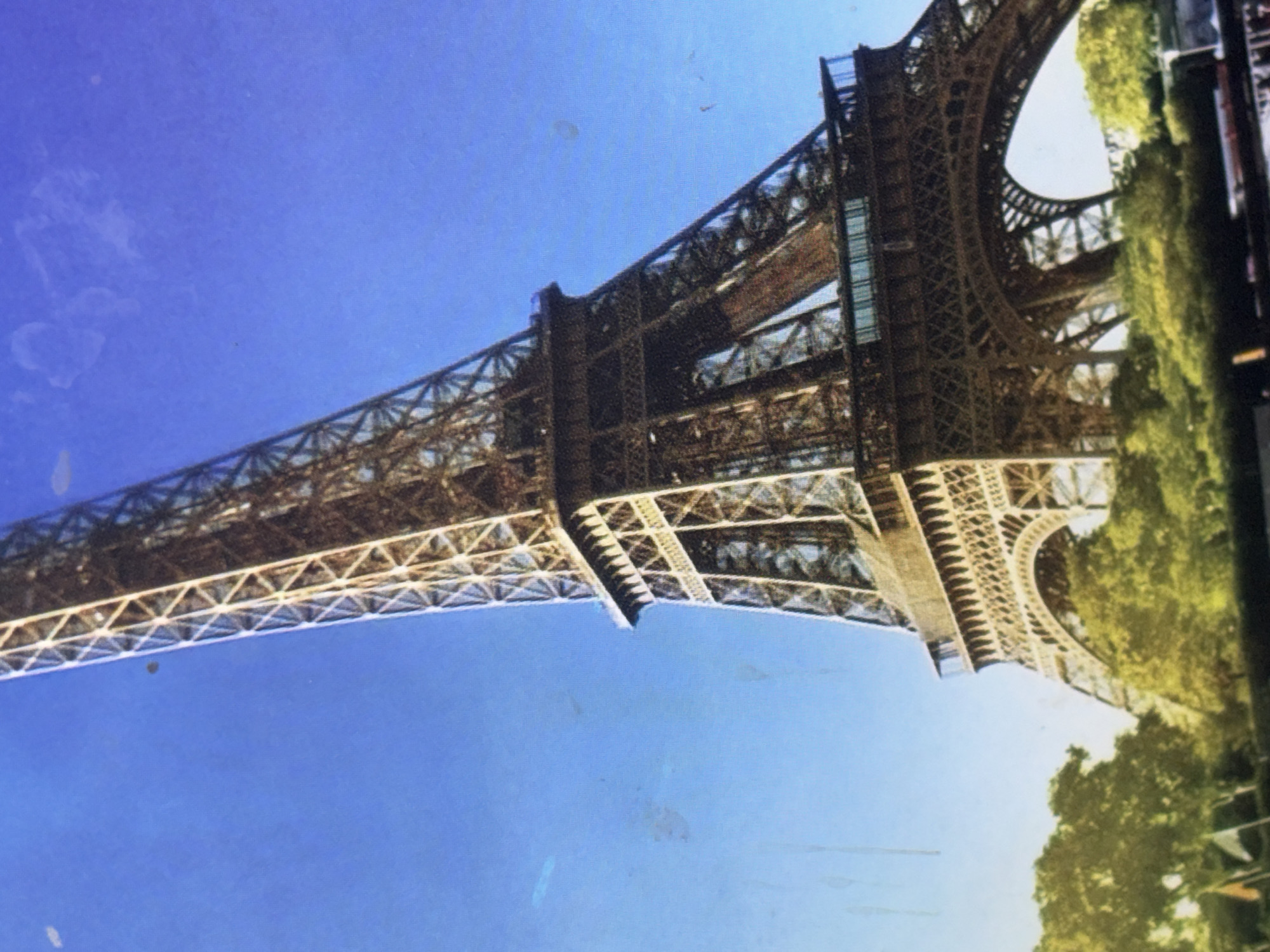The image shows a low-angle, upward view of the Eiffel Tower against a clear blue sky. The iconic iron lattice structure of the tower dominates the frame, with its intricate metalwork visible. Below the tower, lush green trees and foliage are visible, suggesting a park or garden setting. Some faint smudges and dust marks are present on the image. The scene is static and depicts the exterior of the Eiffel Tower. The location of the Eiffel Tower is Paris, France, not Trudovets, Bulgaria.