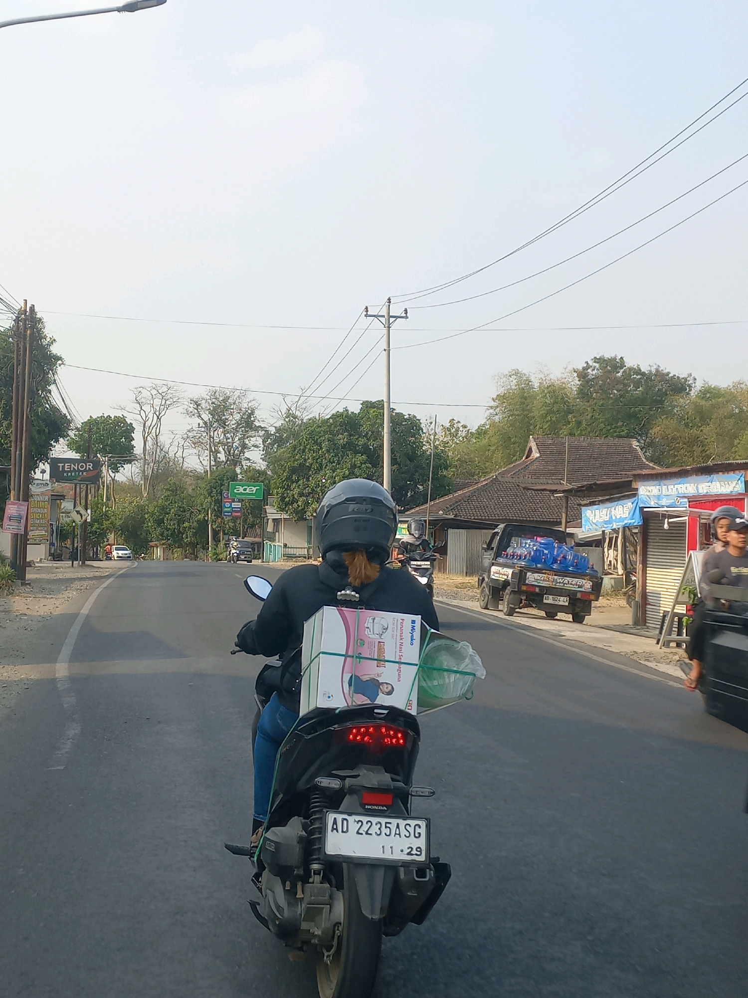 The image captures a street scene in Baturetno, Indonesia, looking forward from the perspective of a motorcycle rider. The road is paved, and on either side, there are buildings and trees. A person wearing a black helmet and a dark jacket is riding the motorcycle, with a large white box secured to the back. The box has green straps and appears to contain goods. A license plate on the motorcycle reads "AD 2235 ASG 11.29".
In the background, a black truck loaded with blue water bottles is visible driving away. On the right side of the road, there's a building with a blue sign that says "MINUMAN SEGAR" and another with "NASI NASI". On the left side, a sign reads "TENOR KRETEK". Overhead power lines are present. The sky is hazy and overcast. The overall atmosphere suggests a rural or semi-urban environment during daylight.