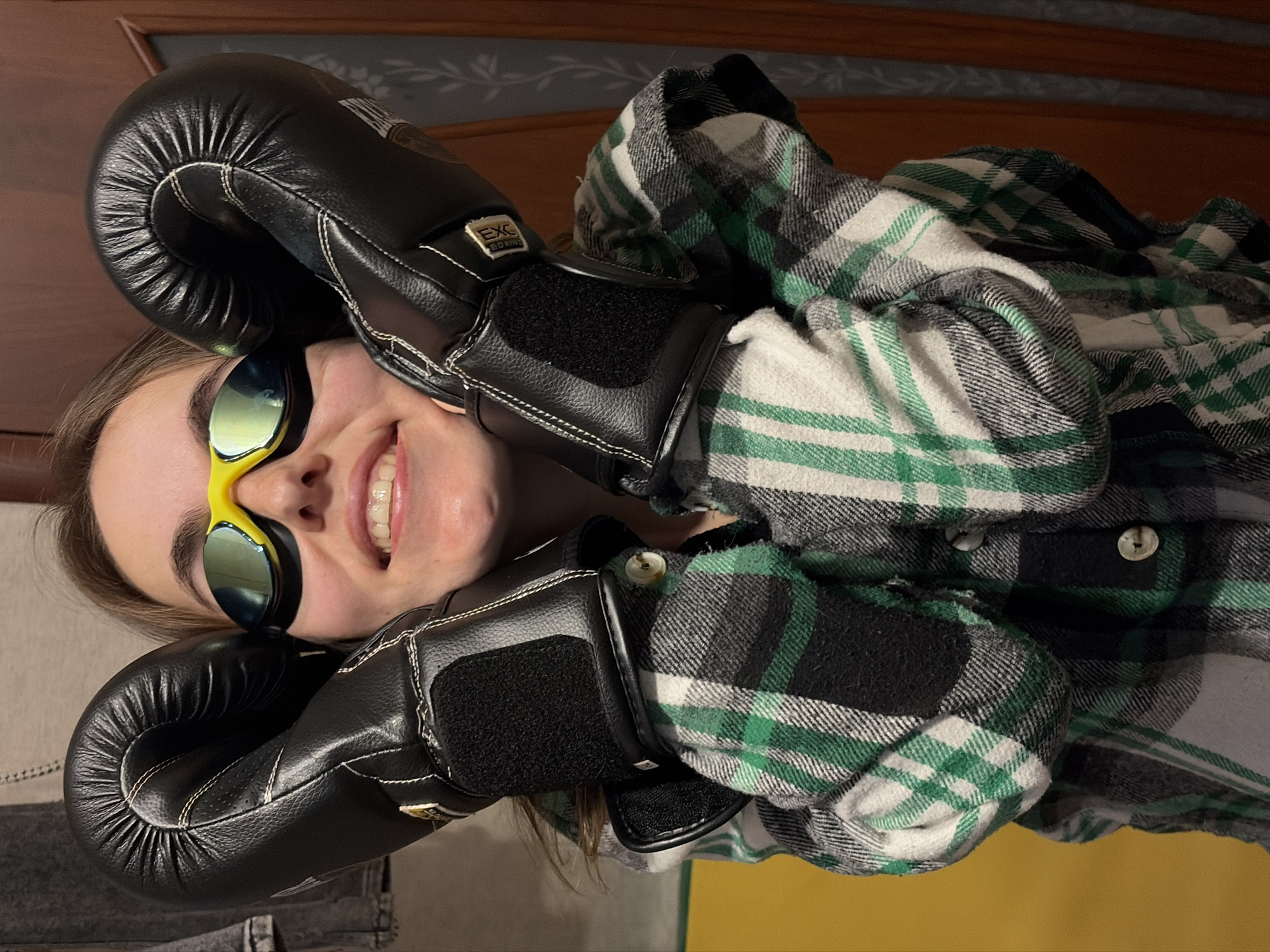 A person is lying on their back, wearing a green and white plaid shirt, black boxing gloves, and swimming goggles. The background includes a brown wooden headboard. The location, Izhevsk, Russia, is not discernible from the visual information provided.