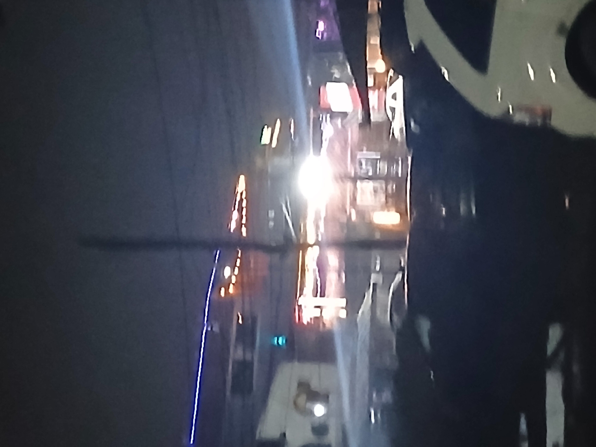 The image depicts a nighttime street scene in Lagos, Nigeria. Streetlights and neon signs illuminate buildings lining the road. A utility pole extends vertically from the left side of the frame. Reflections of lights are visible on the wet street. A vehicle is partially visible in the foreground on the right. Silhouetted figures appear to be walking on the sidewalk.