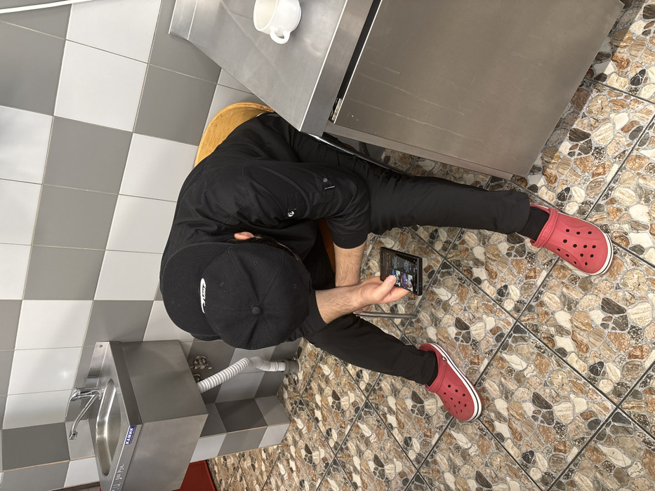 An individual is depicted seated indoors in what appears to be a utility or backroom area within Kirkkolakhti, Russia. The person, a male, is wearing a black long-sleeved work jacket, black pants, a black baseball cap worn backward, and red Crocs with white soles. He is positioned with his head bowed, leaning forward, and holding a dark-colored smartphone horizontally in his left hand, observing its illuminated screen. He is seated on a light brown wooden stool, partially visible behind him. The floor is covered with patterned tiles in shades of brown, beige, and dark grey. The walls are tiled with a checkerboard pattern of alternating grey and white square tiles. To the left, a wall-mounted stainless steel utility sink with a faucet is installed, featuring the "КАМИК" (KAMIK) brand name. A white flexible drain hose extends from beneath the sink. A segment of red flooring or a mat is visible in the lower left corner. To the right, a stainless steel countertop and cabinet unit are present, with a white ceramic mug resting on the countertop.