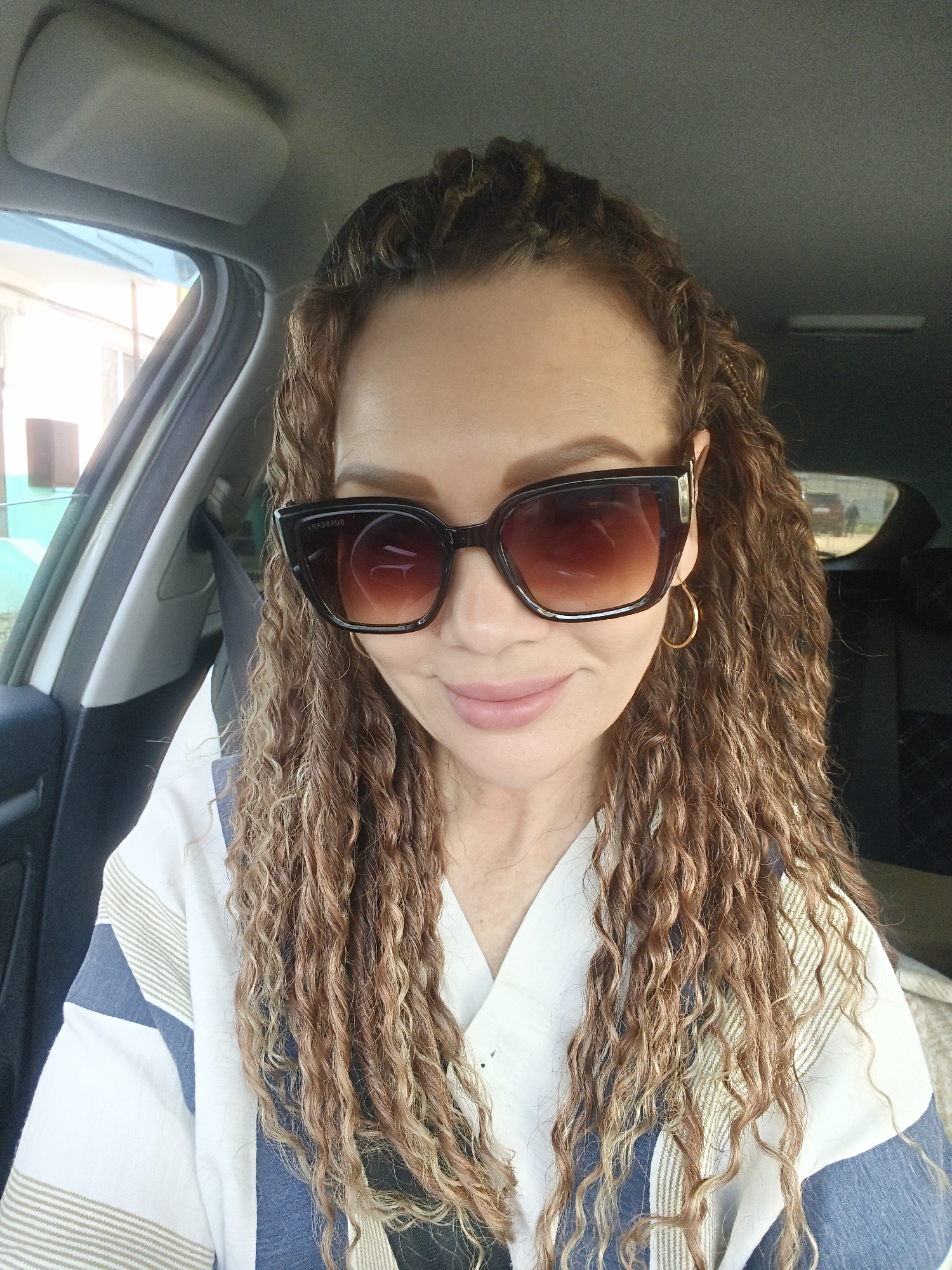 A light-skinned woman with brown, curly hair and sunglasses is seated in the driver's seat of a car. She is wearing a white shirt with blue and tan stripes. The car's interior is visible, including the seatbelt, dashboard, and window. The woman is looking down and smiling slightly. The location appears to be Unknown, Unknown.