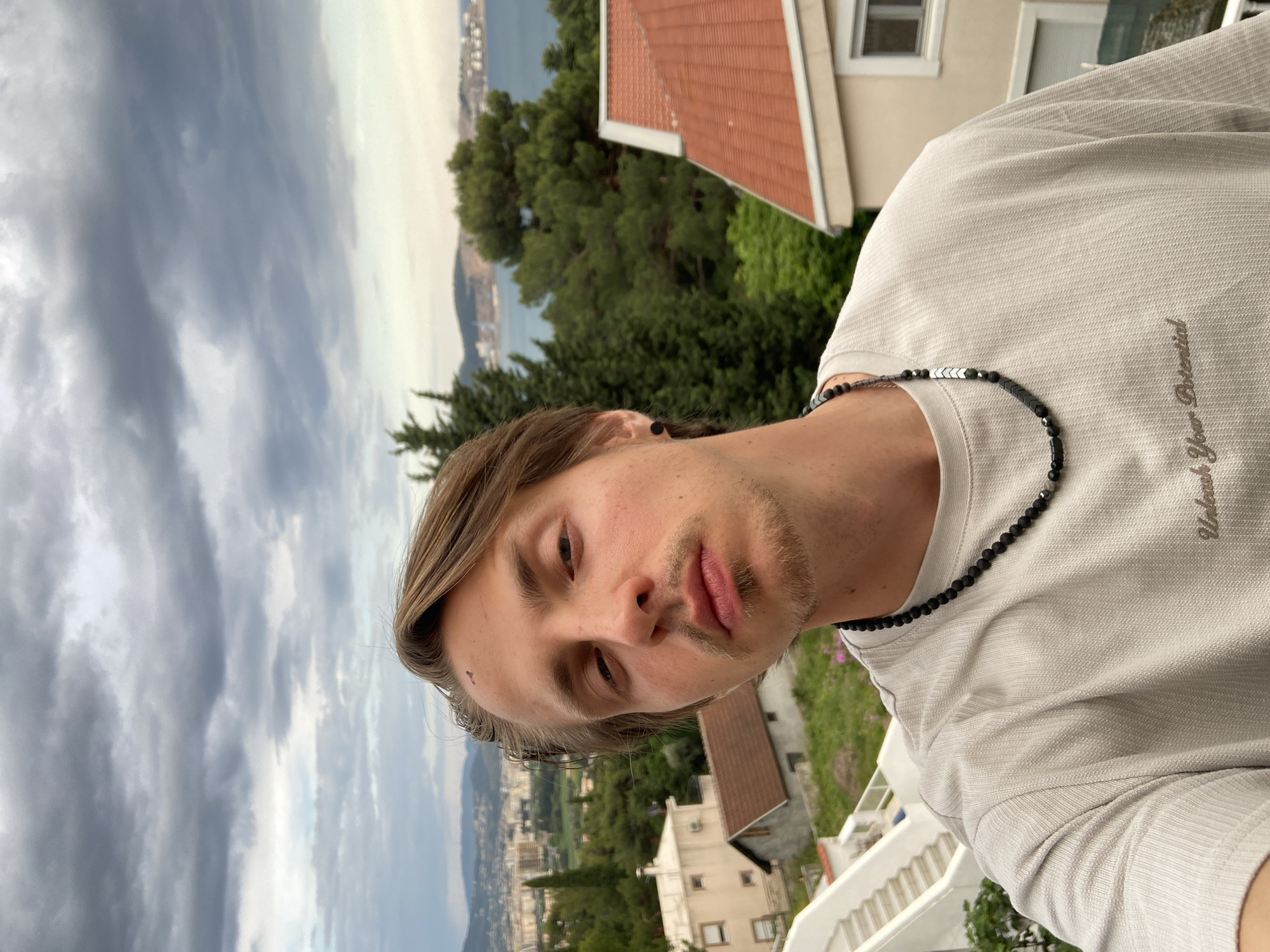A man with light brown hair and a beard is posing for a selfie. He is wearing a light gray shirt with a black bead necklace. The shirt has the words "Unleash Your Potential" stitched into the upper right side of the chest. In the background, there is a view of a city nestled between green trees and the sea, under a cloudy sky. The background suggests a location with a coastal view. Structures like houses with red tile roofs and white walls are also visible. Based on the location suggested and the presence of typical Montenegrin architecture, it is plausible that the image was taken in Šušanj, Montenegro, a coastal town with similar characteristics.