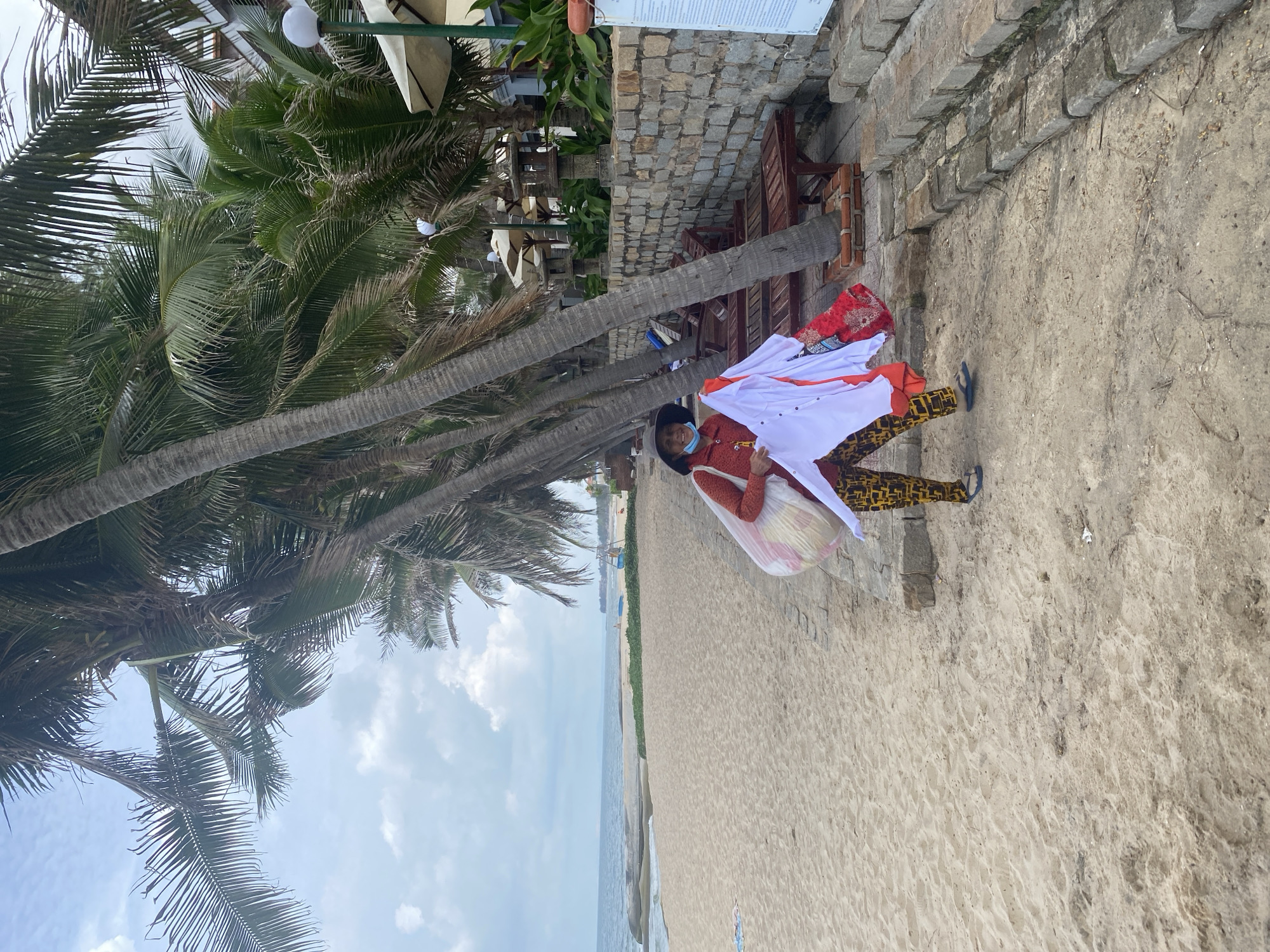 A woman wearing a face mask, a red top, and patterned pants stands on a sandy beach in Phu Thuy Ward, Vietnam. She is holding a large, translucent bag filled with what appear to be folded clothes. In her right hand, she holds up a white shirt and a red patterned garment. The beach has a line of palm trees, lounge chairs, and a stone wall behind her. The ocean is visible in the background. The sky is partly cloudy.