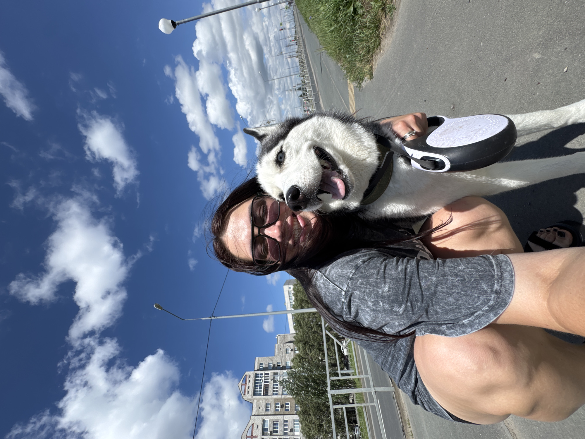 A woman and a husky dog are taking a selfie outdoors on a sunny day. The woman is wearing sunglasses and a grey t-shirt, crouching down to be at the dog's level. She has long dark hair that is blowing in the wind. The husky is smiling with its tongue out, its blue eyes clearly visible. In the background, there are apartment buildings, trees, a street, a sidewalk, and a cloudy blue sky. The image appears to be taken on a street in Tolyatti, Russia, likely near a residential area given the presence of apartment buildings. The woman is holding a retractable dog leash.