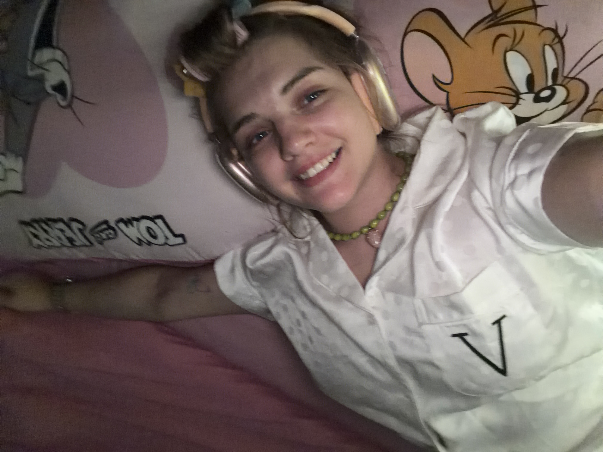 A young woman with fair skin, blue eyes, and a bright smile lies in bed. She is wearing gold headphones and a white, short-sleeved pajama top with a black "V" on the pocket. She also wears a green beaded necklace. She is positioned between two pink Tom and Jerry-themed pillows. The scene seems to be indoors, possibly in Большая Шильна, Russia, but there are no location-specific details in the image to confirm. The subject is taking a selfie.