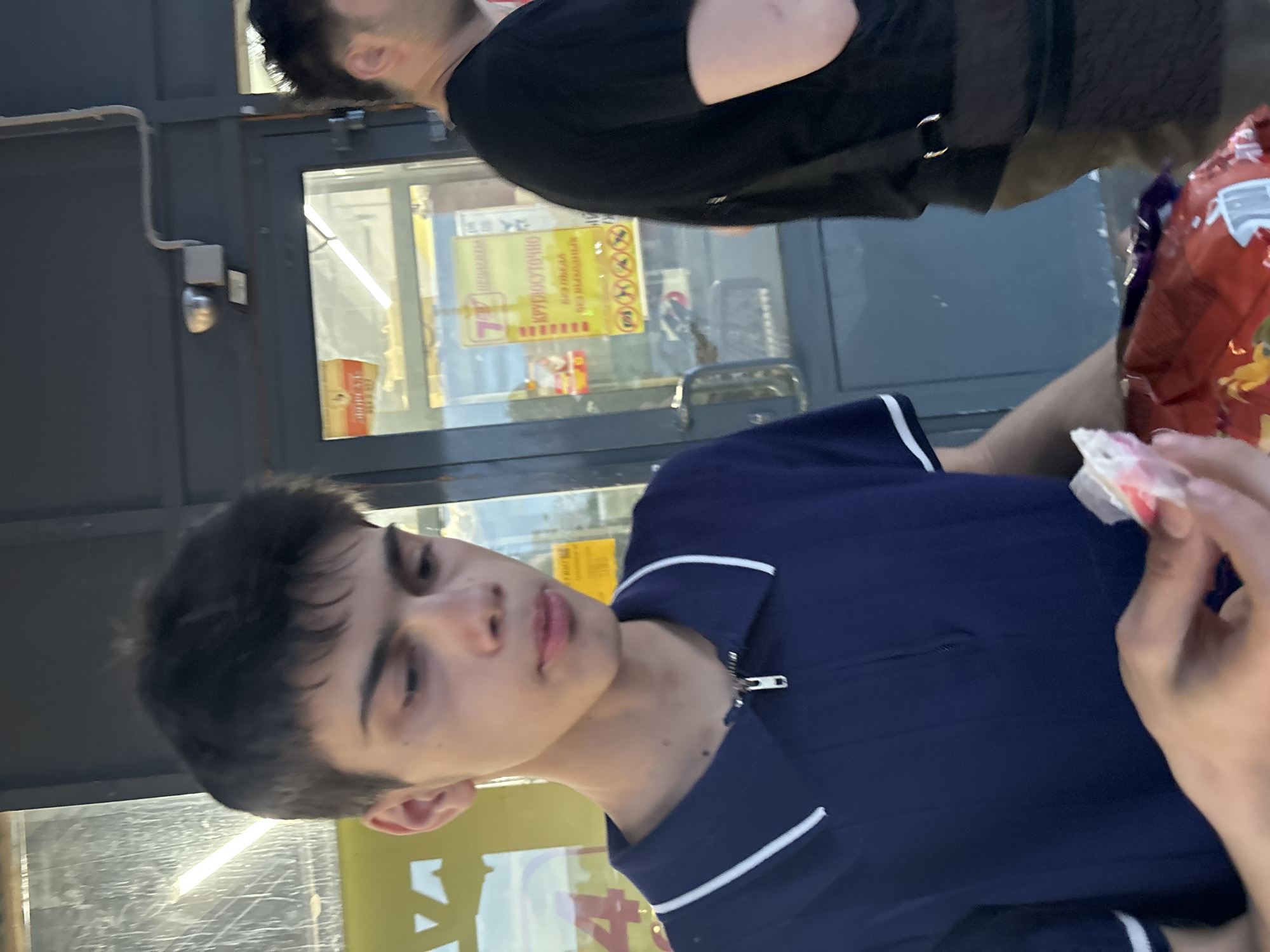 A young male with dark hair and fair skin is pictured outdoors, wearing a navy blue polo shirt with white trim. He is holding a bag of chips in his right hand and a small white object in his left hand, which he appears to be inspecting. His gaze is directed downwards. Behind him, a glass-paned door leads into a building with fluorescent lighting visible inside. A sign on the door displays text in Russian, including the words "КРУГЛОСУТОЧНО" (around the clock). Another person, a male with dark hair wearing a black t-shirt, is partially visible to the right, with a bag slung over his shoulder. The scene suggests an urban setting, possibly in front of a store or commercial establishment.
