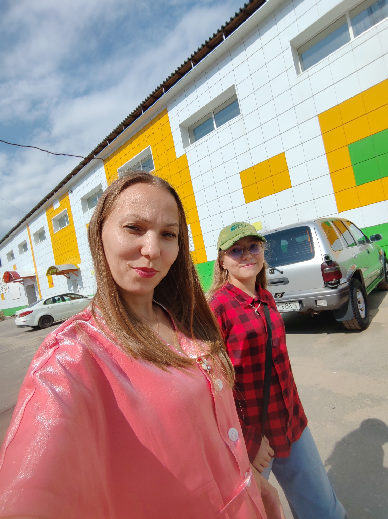 In Syktyvkar, Russia, two individuals are captured in a selfie with a building and vehicles in the background. The person in the foreground, on the left, is a woman with long, blonde hair and is wearing a shiny pink shirt. She is looking towards the camera with a slight smile. To her right and slightly behind her is a younger individual, possibly a girl, wearing a green cap, round sunglasses, and a red and black plaid shirt. They are also looking towards the camera with a smile.
The building behind them has a distinctive facade with alternating white, yellow, and green square panels. Several windows are visible on the building. In the middle ground, a silver SUV is parked, and to the left, a white sedan is also visible. The ground appears to be asphalt or a similar paved surface. The sky above is blue with scattered white clouds, indicating daytime. The overall scene suggests a casual outdoor setting in an urban or semi-urban environment.