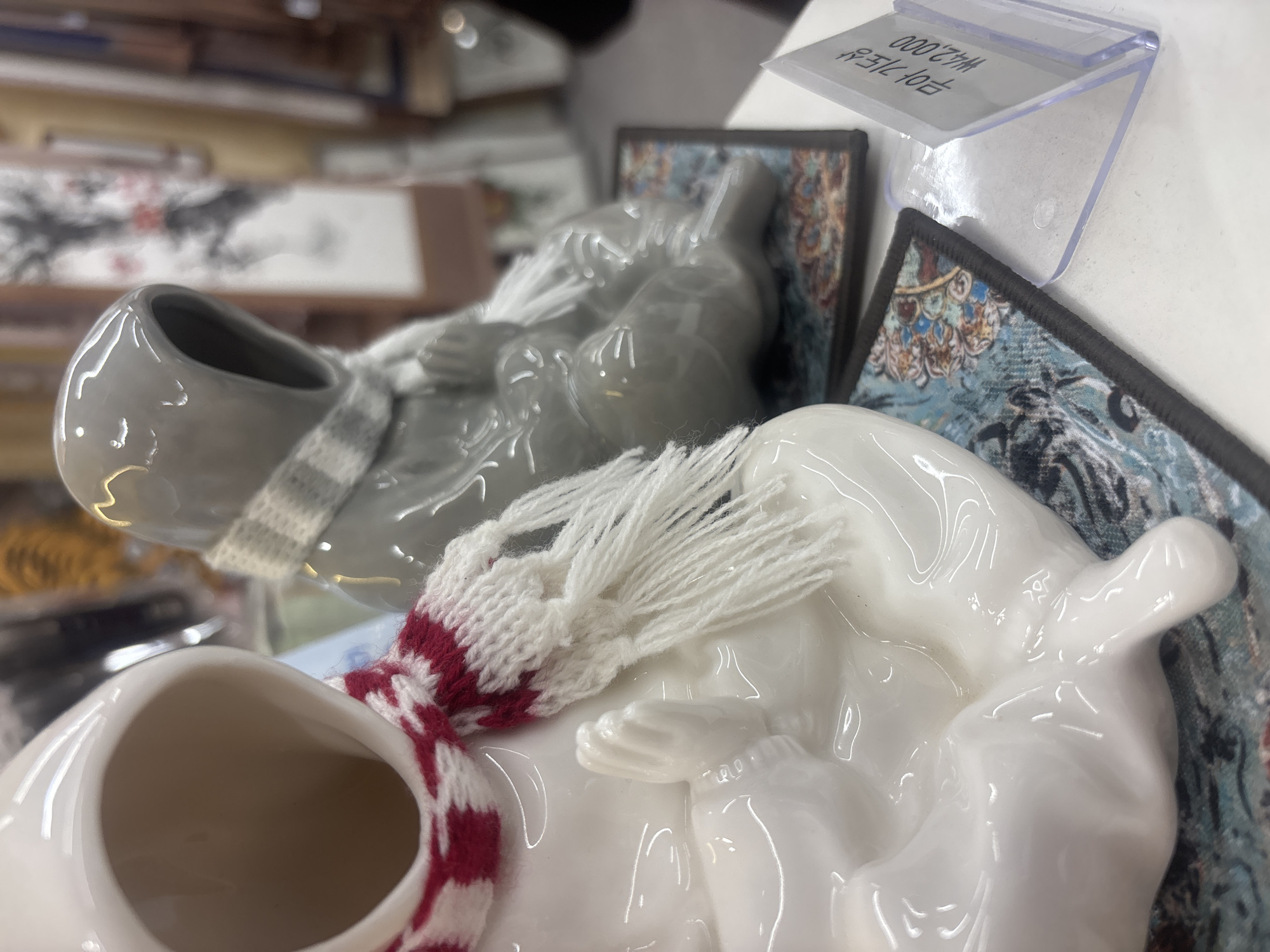 Two ceramic figures, one white and one gray, are seated side-by-side on decorative mats. The figures have hooded heads and appear to be wearing scarves. The white figure has a red and white striped scarf with white fringe, while the gray figure has a gray and white checkered scarf. The white figure's hands are clasped in front of its body. Both figures are positioned on what appears to be a white surface, possibly a table or shelf. In the foreground, a clear plastic price tag holder is visible, displaying Korean text and the price "₩42,000". In the background, out-of-focus items are visible, including what appear to be more decorative objects and possibly some artwork. The overall impression is that of items displayed in a shop or market setting. The presence of the price tag in Korean indicates this is likely in South Korea, and given the context of souvenir shops and cultural items, a location in Gyeongju-si, a city known for its historical and cultural significance in South Korea, is plausible.