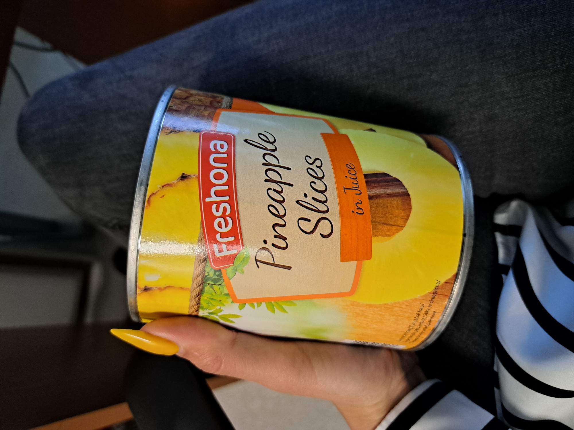 A hand with a yellow fingernail is holding a can of Freshona brand pineapple slices in juice. The can is prominently displayed, and its label reads "Pineapple Slices in Juice" in a brown font. The label also includes the Freshona logo in red and green. The background includes a person wearing dark denim pants and a black and white striped garment. The location is not discernible in the image.