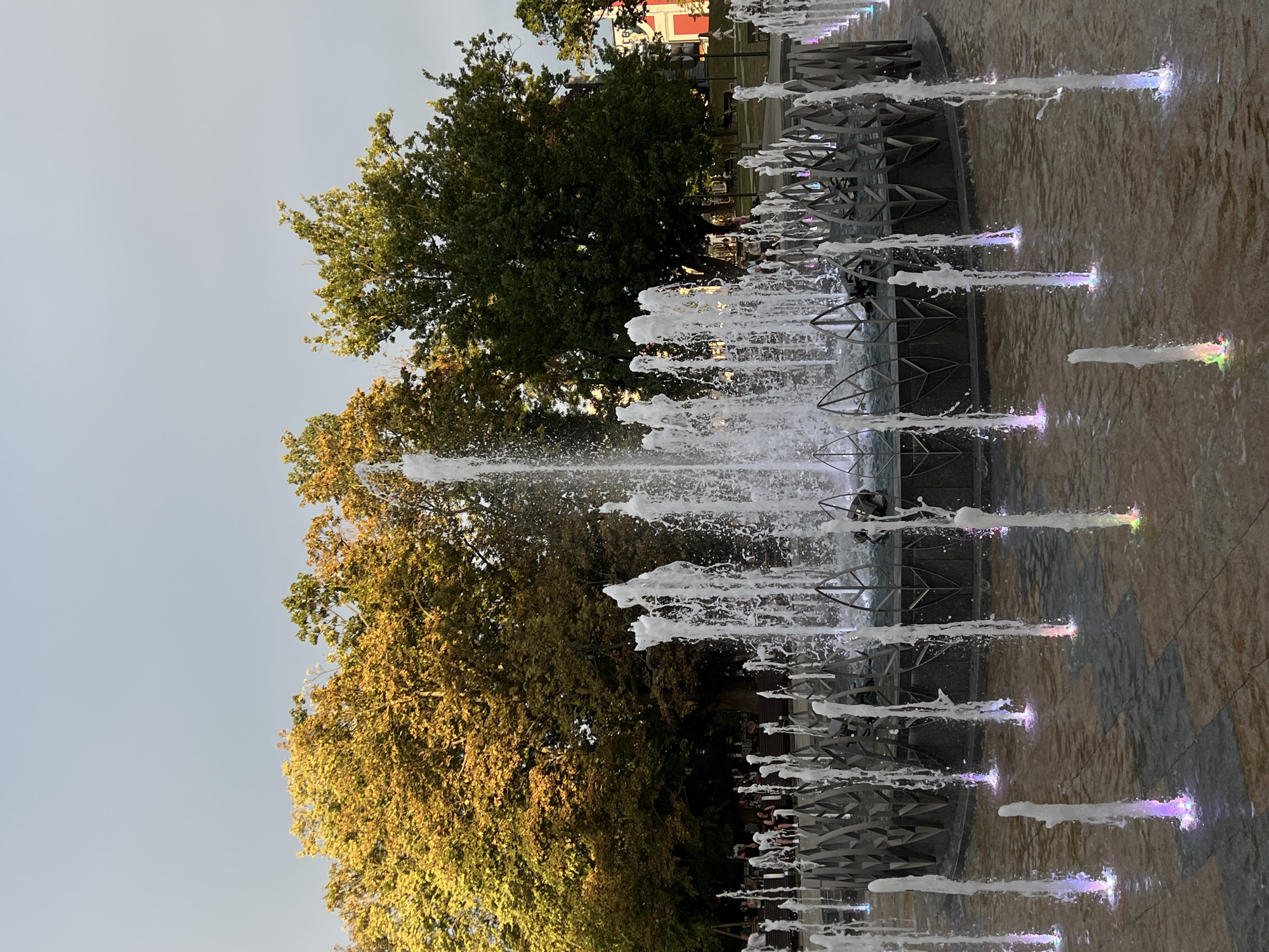 The image shows a public fountain with multiple jets of water in motion, illuminated by colored lights at their base. The fountain is located in a paved area, likely a square or park, with trees in the background. Some trees on the left have yellow and orange foliage, suggesting it may be autumn. Several people are visible in the background, seated on benches or walking. In the far background, a building with red and white accents is partially visible. The overall scene depicts a serene outdoor setting in what is identified as the city of Lublin, Poland.