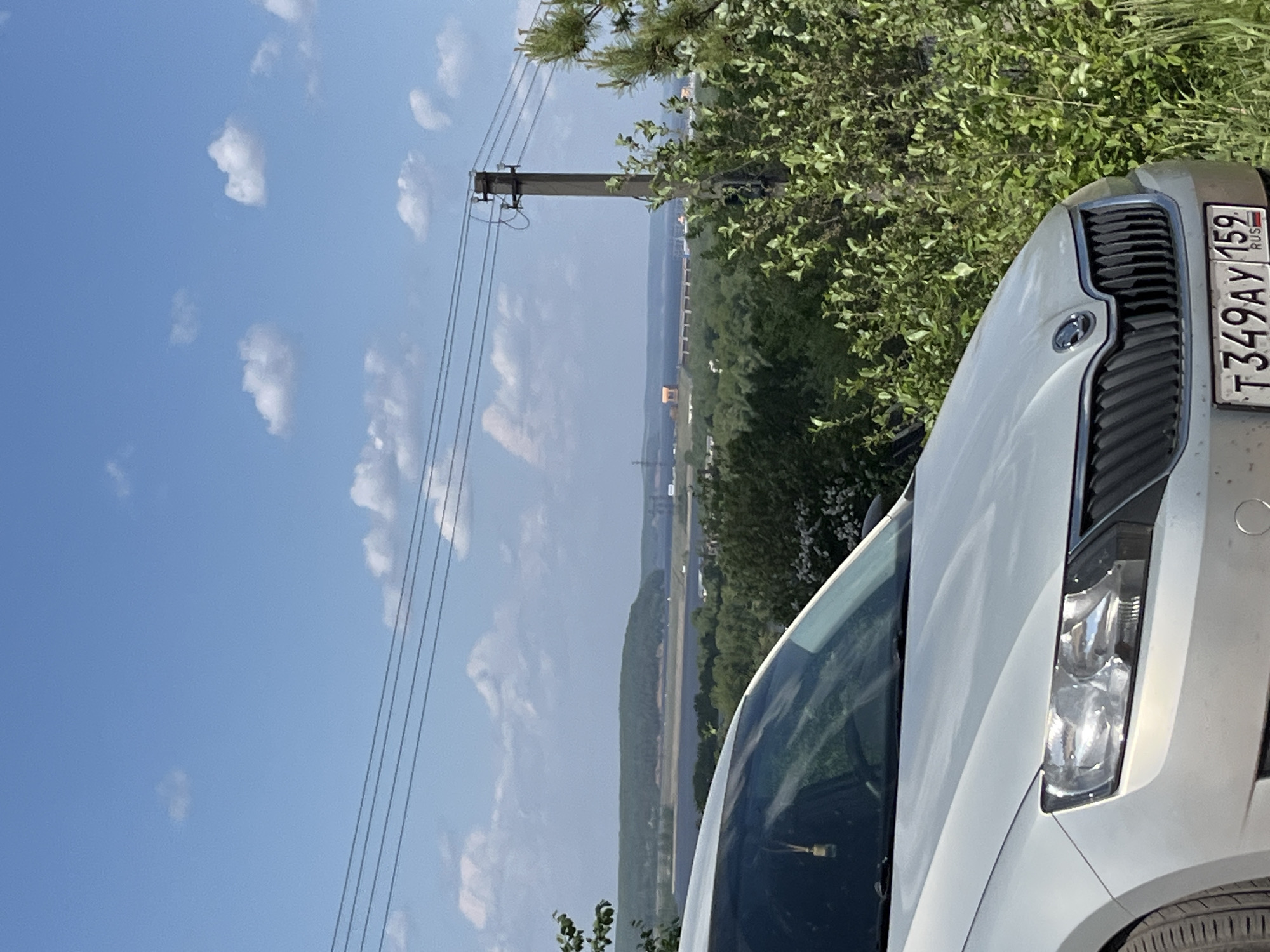 The image shows a view in the city of Chaikovsky, Russia. Part of a silver Skoda car is in the foreground, its front grill and headlight visible, along with the license plate reading "T349AY 159 RUS". Behind the car, a landscape extends into the distance. The sky is blue with scattered white clouds. Electrical lines run from a utility pole towards the distant horizon. A green area with trees and vegetation partially obscures the middle ground. Further in the background, there is a landscape with what appears to be a river or large body of water, and the faint outline of buildings in the distance. The overall scene gives the impression of a rural or suburban area.