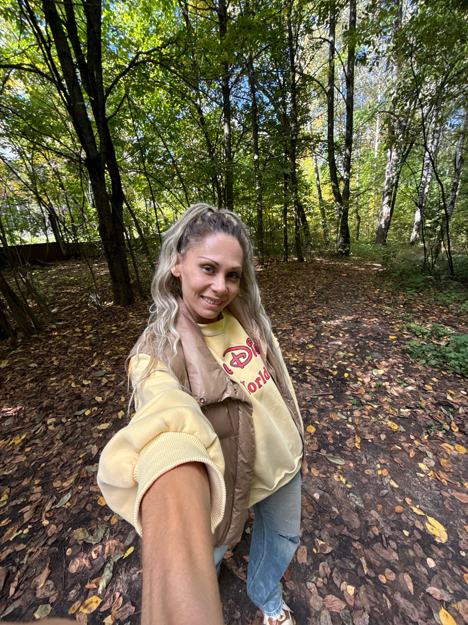 A woman is taking a selfie in an autumnal forest path in Rusanovo, Russia. She has long blonde hair pulled into a ponytail, is wearing a light yellow sweatshirt with a graphic print, a brown vest, and ripped blue jeans. The ground is covered in fallen leaves.