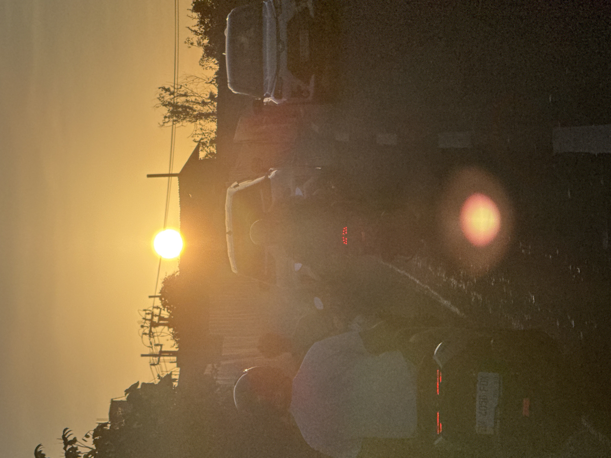 At dusk, Ungasan, Indonesia, a sun glare obscures details on a road lined with utility poles and vegetation on the left. A group of people, obscured by sunlight, are present. Vehicles, including motorcycles and what appears to be a truck, are also on the road. One of the motorcycles has a visible license plate.