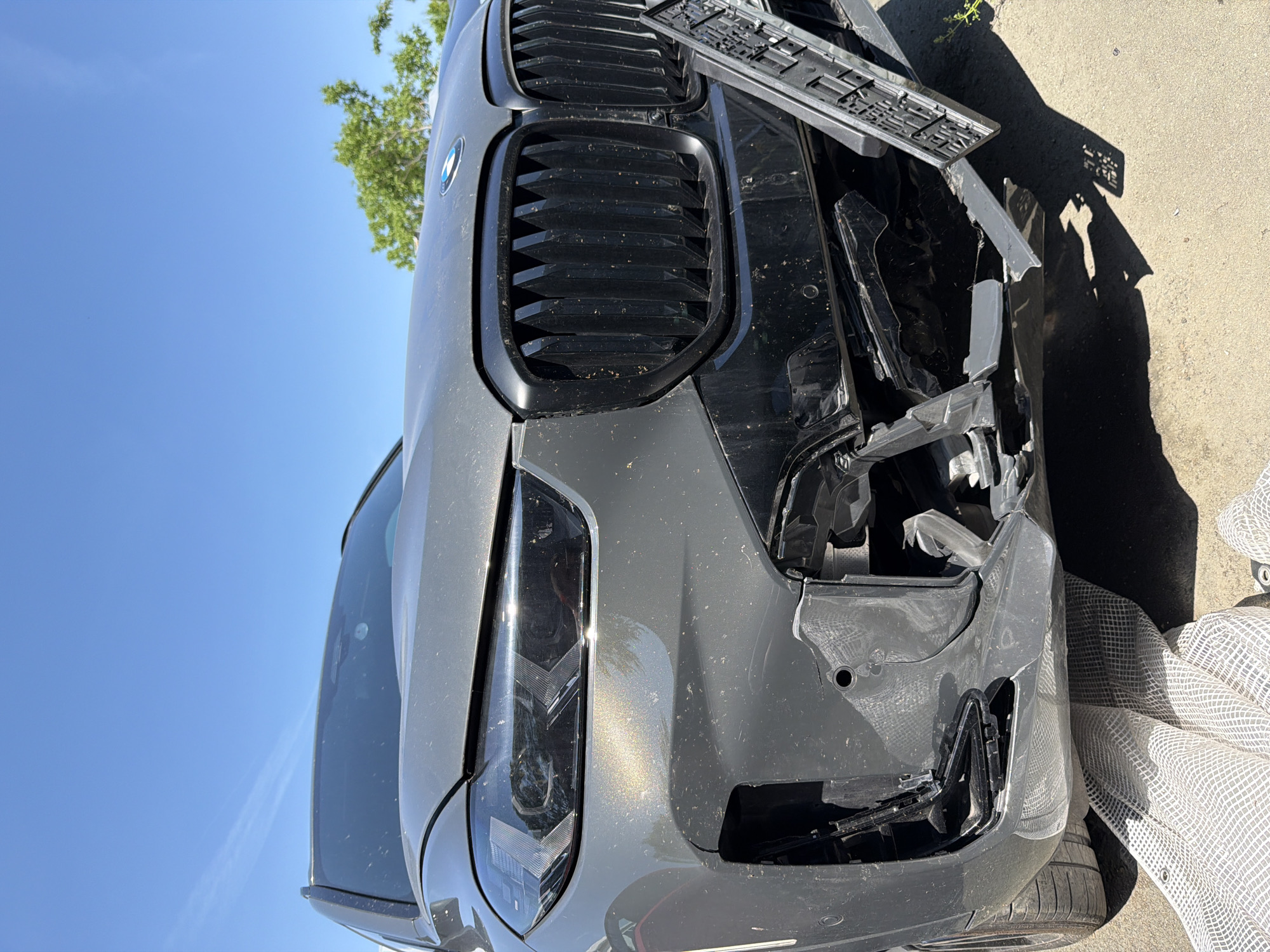 The image shows the front of a gray BMW with significant damage to its front end. The bumper is partially detached and cracked, with internal components exposed. The front grill is intact but dirty. The driver's side headlight appears undamaged. A license plate holder lies displaced on top of the car. The car is located outdoors on a paved surface. The sky is blue with some green foliage visible in the background. There is no evidence to suggest that the image was taken in Burgas, Bulgaria.