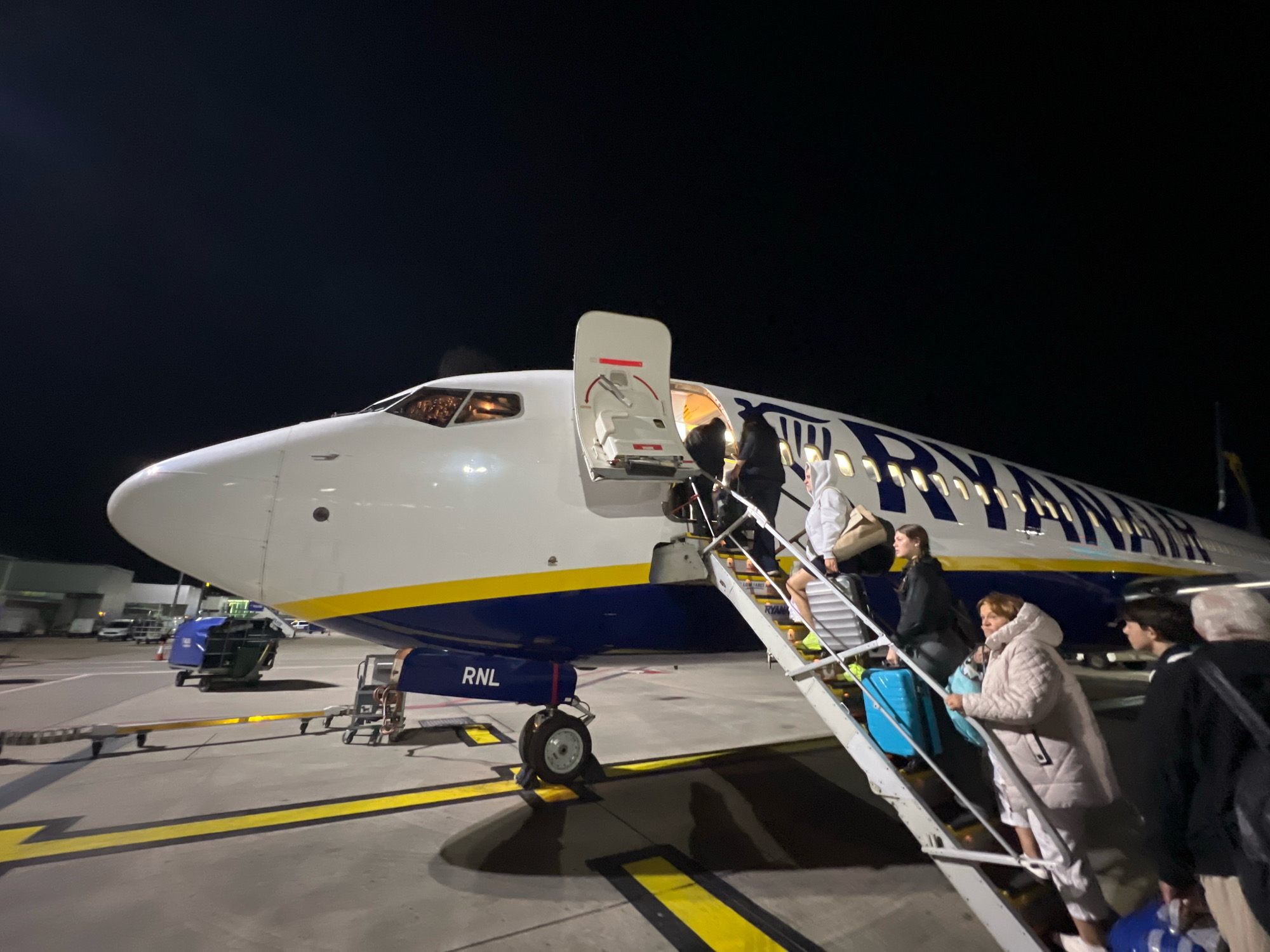 In Uttlesford, United Kingdom, at night, passengers board a Ryanair airplane via mobile stairs. The aircraft is white with a blue and yellow livery, featuring the Ryanair logo on its fuselage. Several people are visible on the stairs, carrying luggage, and moving towards the aircraft door, which is open. The scene is illuminated by artificial lights, and the dark sky is visible above. The airport tarmac and some airport equipment, including baggage carts, are also present in the foreground and background.