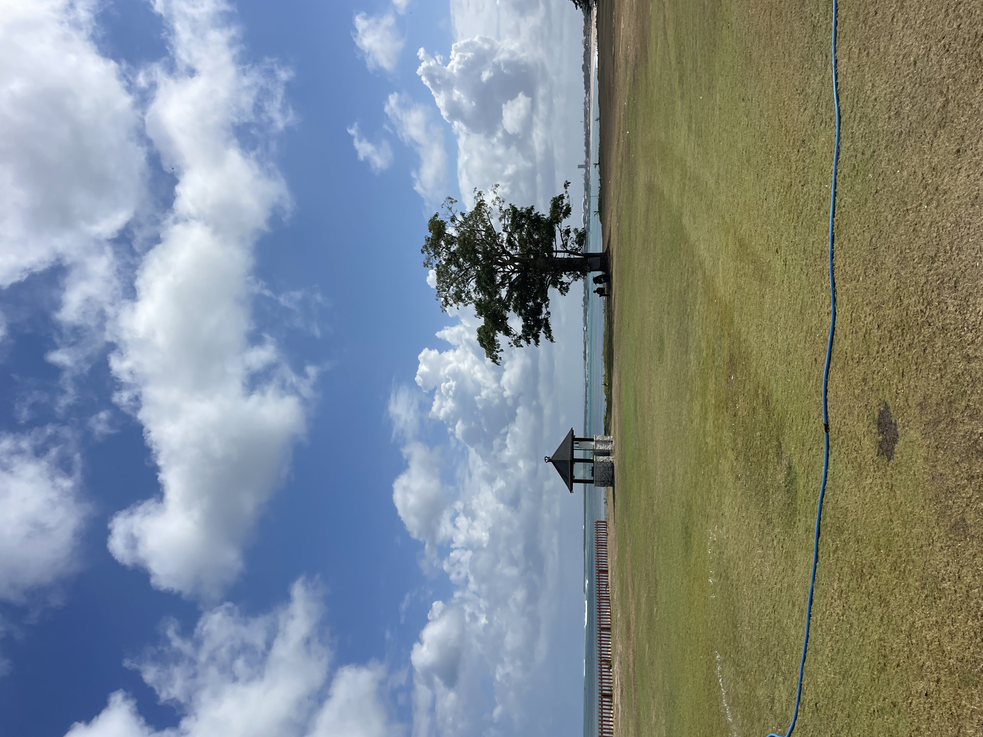 In Kuta Selatan, Indonesia, a wide, grassy field stretches towards a calm ocean under a vibrant blue sky filled with scattered white clouds. In the middle ground of the field, a lone, mature tree stands tall, with two individuals seated beneath its shade. To the left of the tree, a small, stone and dark-roofed gazebo is visible. Further in the distance, a wooden fence runs along the edge of the field, separating it from the ocean. The water appears to have gentle waves. The overall scene depicts a serene outdoor environment.