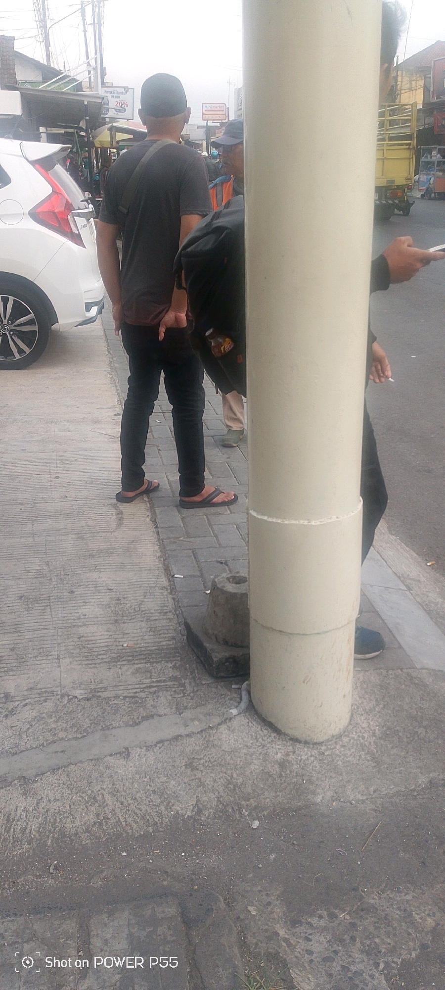 The image captures a street scene in Unknown, Unknown, with a focus on several individuals standing near a large, pale, cylindrical object. The scene appears to be outdoors, possibly a sidewalk or street.
In the foreground, a person wearing dark pants and flip-flops stands with their back to the camera. To their right, another person, partially obscured by the cylindrical object, is seen from the waist down, wearing dark pants and dark shoes. This person is holding a cigarette in their right hand and what appears to be a mobile phone in their left hand, though it is mostly hidden.
Further back, a white car is parked on the left side of the frame. Beyond the car, there are several figures, including one wearing a dark t-shirt and a cap, with a strap across their chest. Another individual is visible with a blue cap and an orange vest. In the background, there are buildings, market stalls, and a yellow truck. The ground is paved with concrete and interlocking bricks. The lighting suggests it is daytime, with overcast skies. A water bottle with a yellow label is partially visible in a bag. A discarded cigarette butt is visible on the ground near the cylindrical object.
