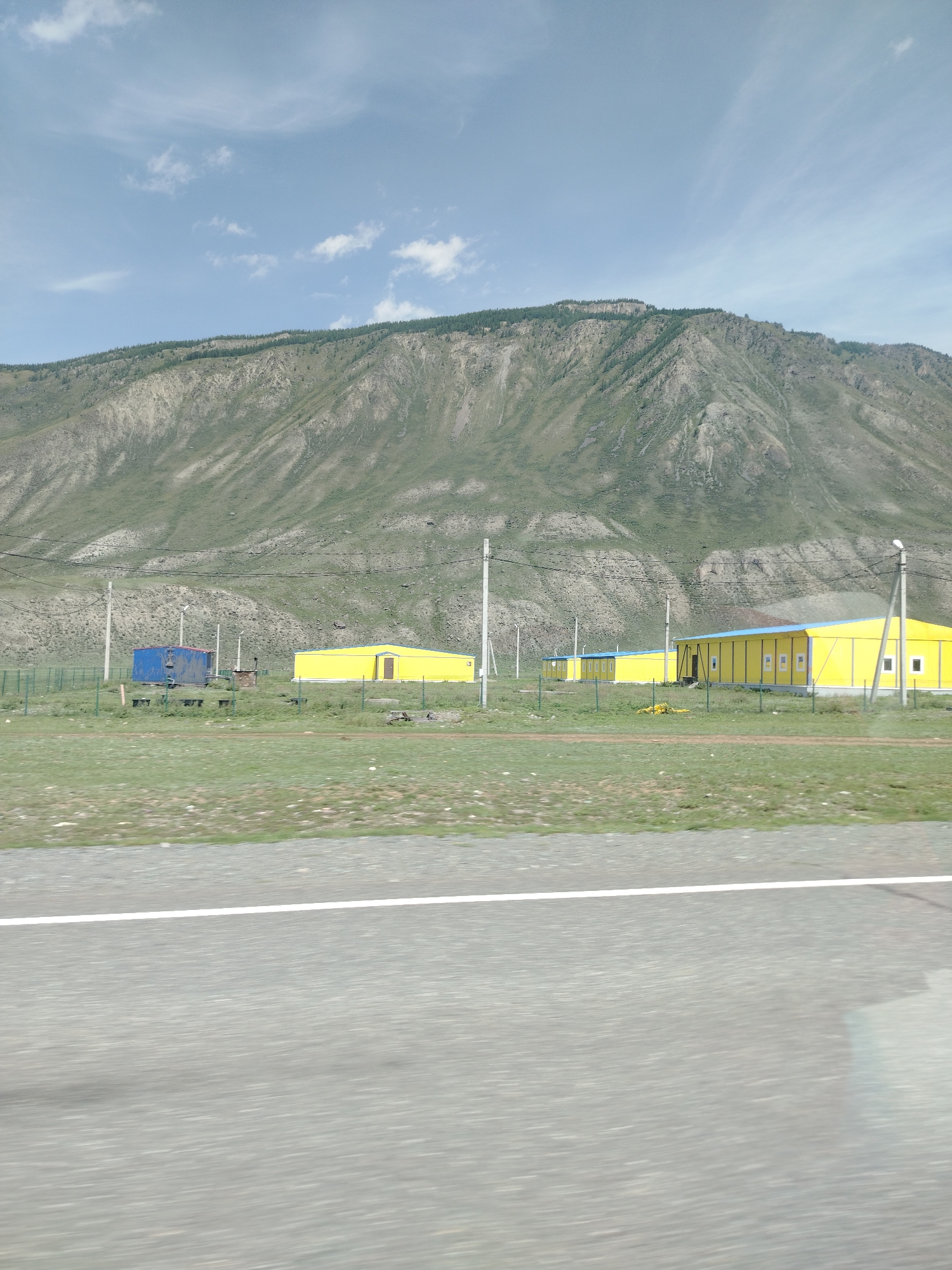 The image shows a view of the village of Chibit, Russia, with a large mountain in the background. In the foreground, there is a road with a white line. Beyond the road, there are several yellow buildings with blue roofs, possibly modular housing or storage units, enclosed by a green fence. A blue building is also visible to the left. Power lines and utility poles are present in the area. The mountain is covered with sparse vegetation and shows signs of erosion. The sky is partly cloudy.