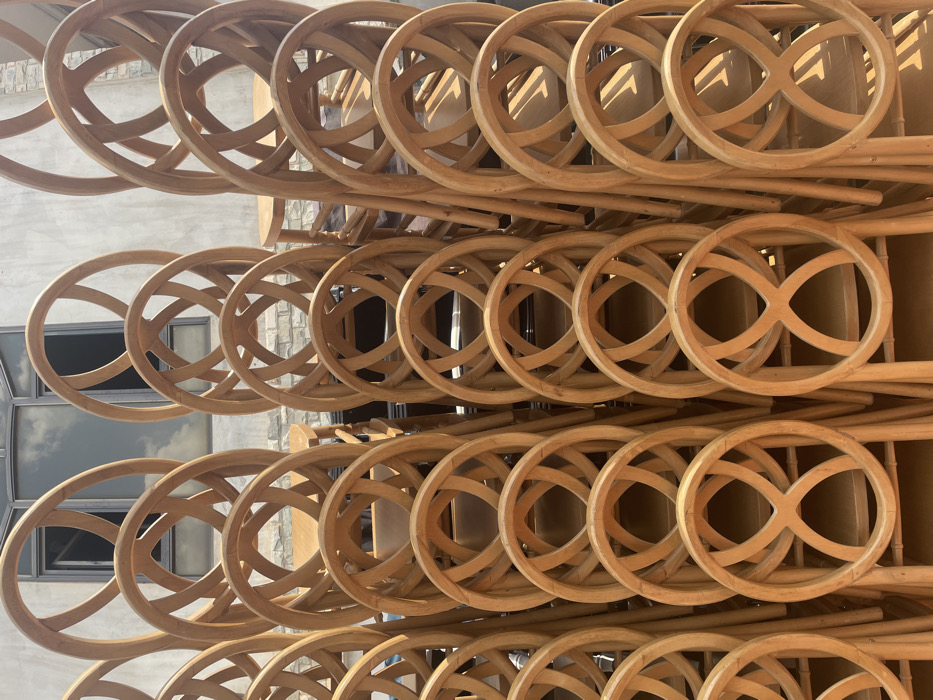 The image depicts a dense arrangement of numerous light-colored wooden chairs, stacked and stored in vertical columns. Each chair features a round backrest with an open, interwoven design resembling a figure-eight. The wood has a natural, light brown hue. In the upper background, a building with a light-colored, possibly stone or brick, facade is visible, featuring large, dark-framed windows reflecting a cloudy sky. The scene is brightly lit by daylight, casting distinct shadows. No individuals are present, and no actions or interactions are occurring. The location is in the city of Unknown, Ghana.